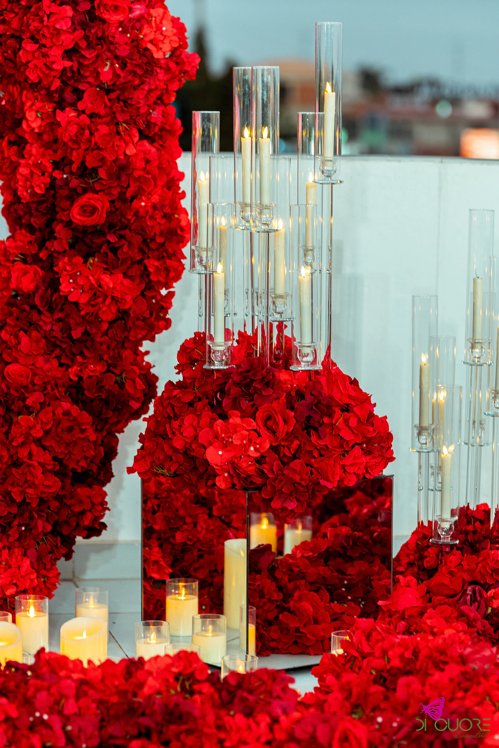 Candelabros de vidro com velas altas sobre um maciço de rosas vermelhas, com um espelho entre as flores