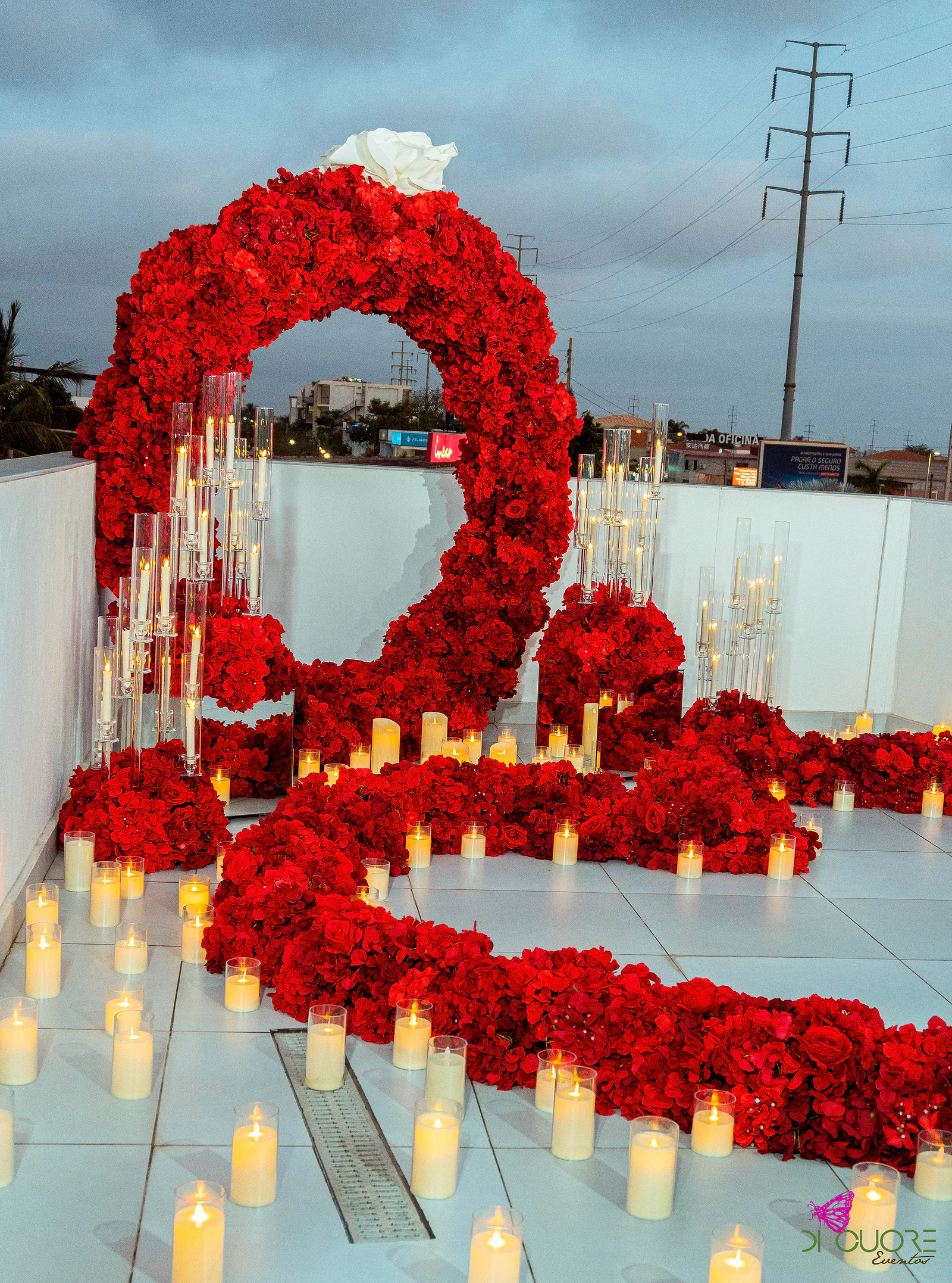 Arco circular de rosas vermelhas ao entardecer, com uma fita de rosas e velas em copos de vidro a avançar sobre o piso claro