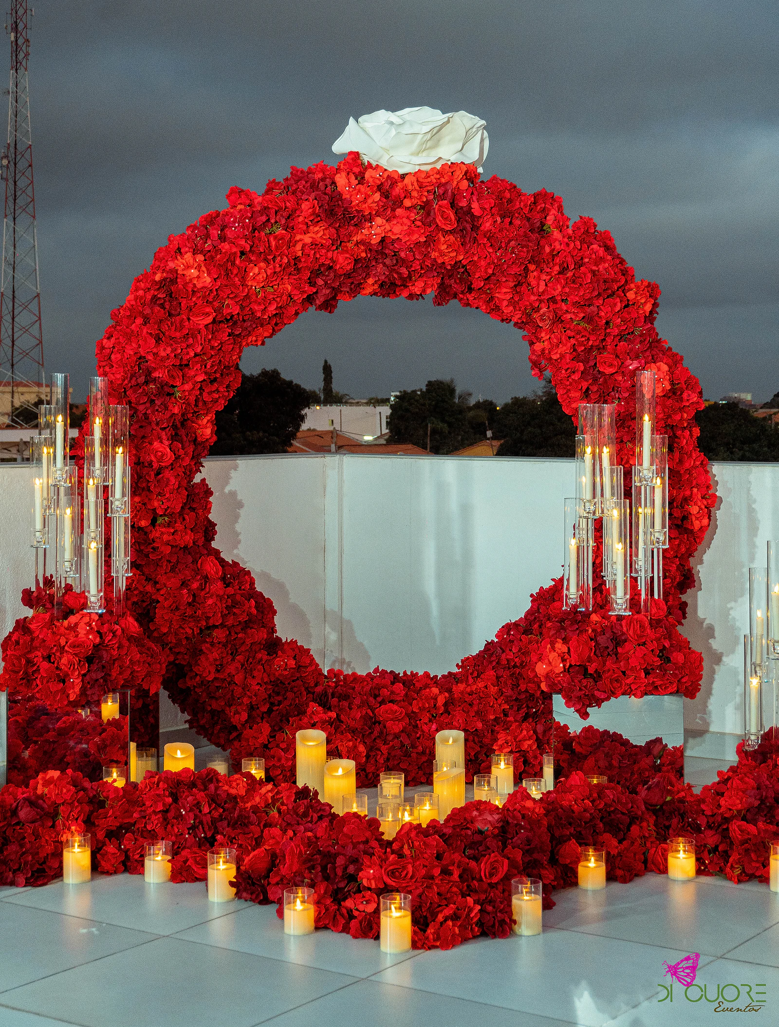 Arco circular de rosas vermelhas com uma rosa branca no topo e candelabros de vidro, rodeado de velas sobre o piso claro, ao entardecer