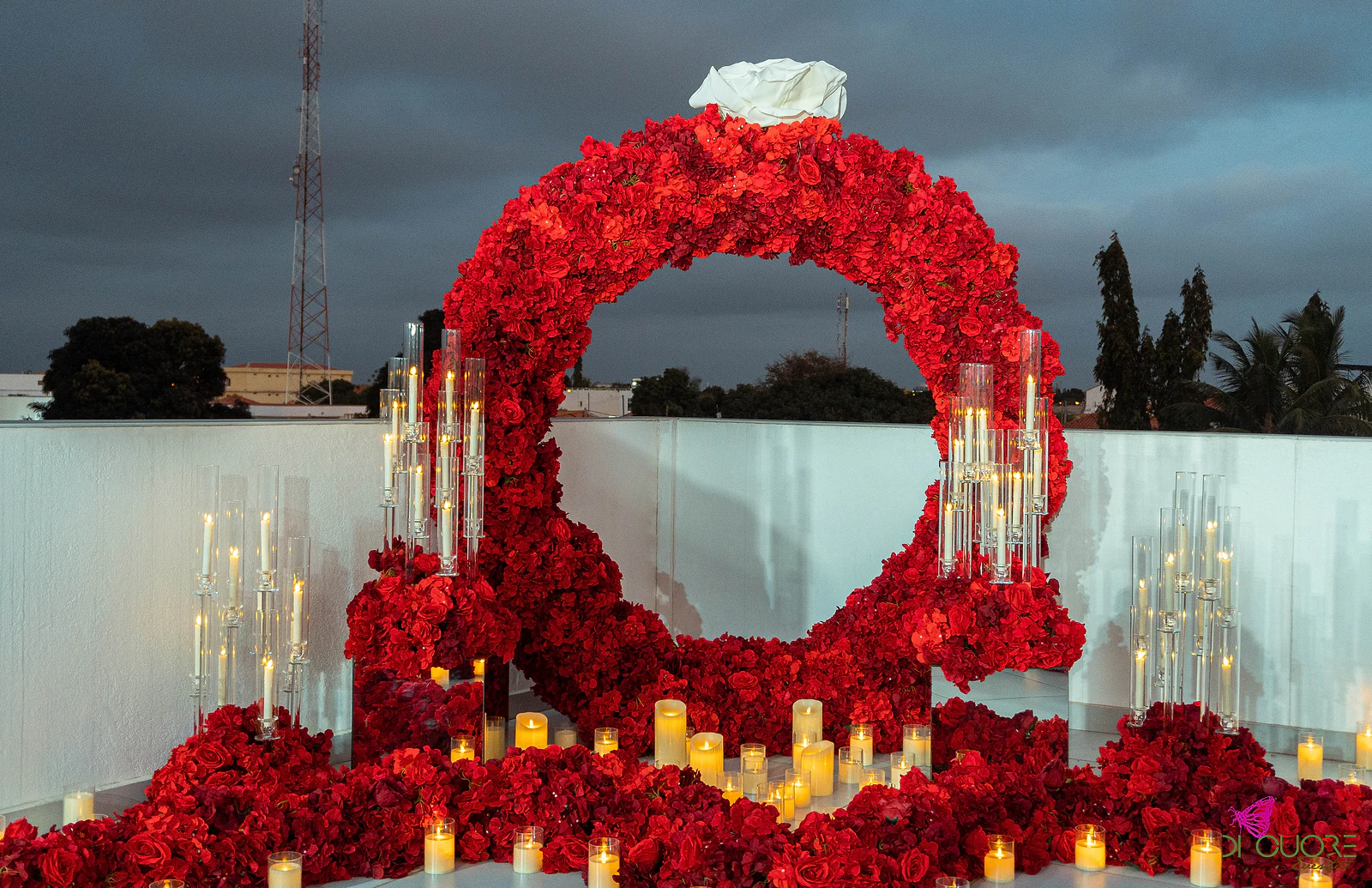 Pormenor do arco de rosas vermelhas com a rosa branca no topo, candelabros de vidro e velas acesas, sob um céu escuro de fim de tarde