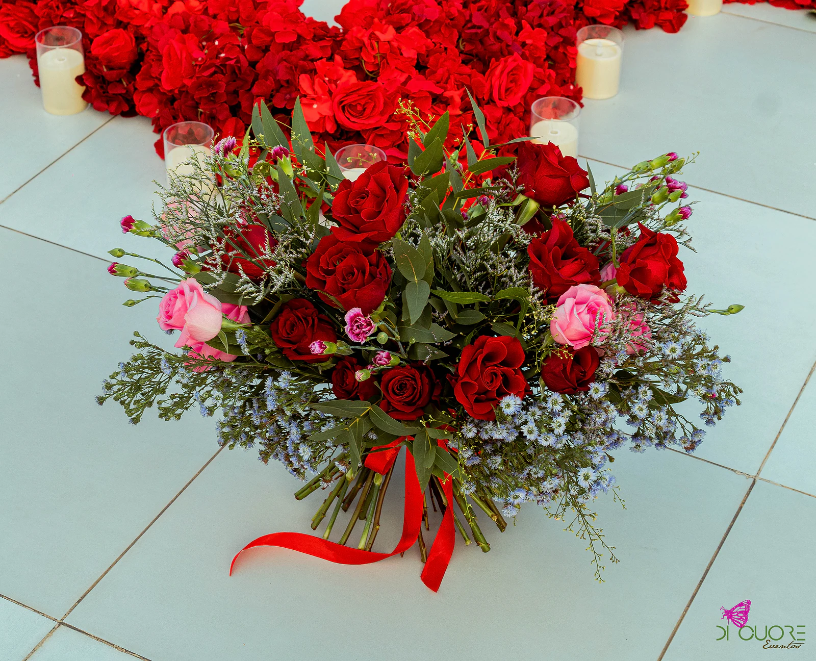 Ramo de rosas vermelhas e cor-de-rosa com flores azuis e fita vermelha, visto de cima sobre o piso claro, com velas e rosas ao fundo