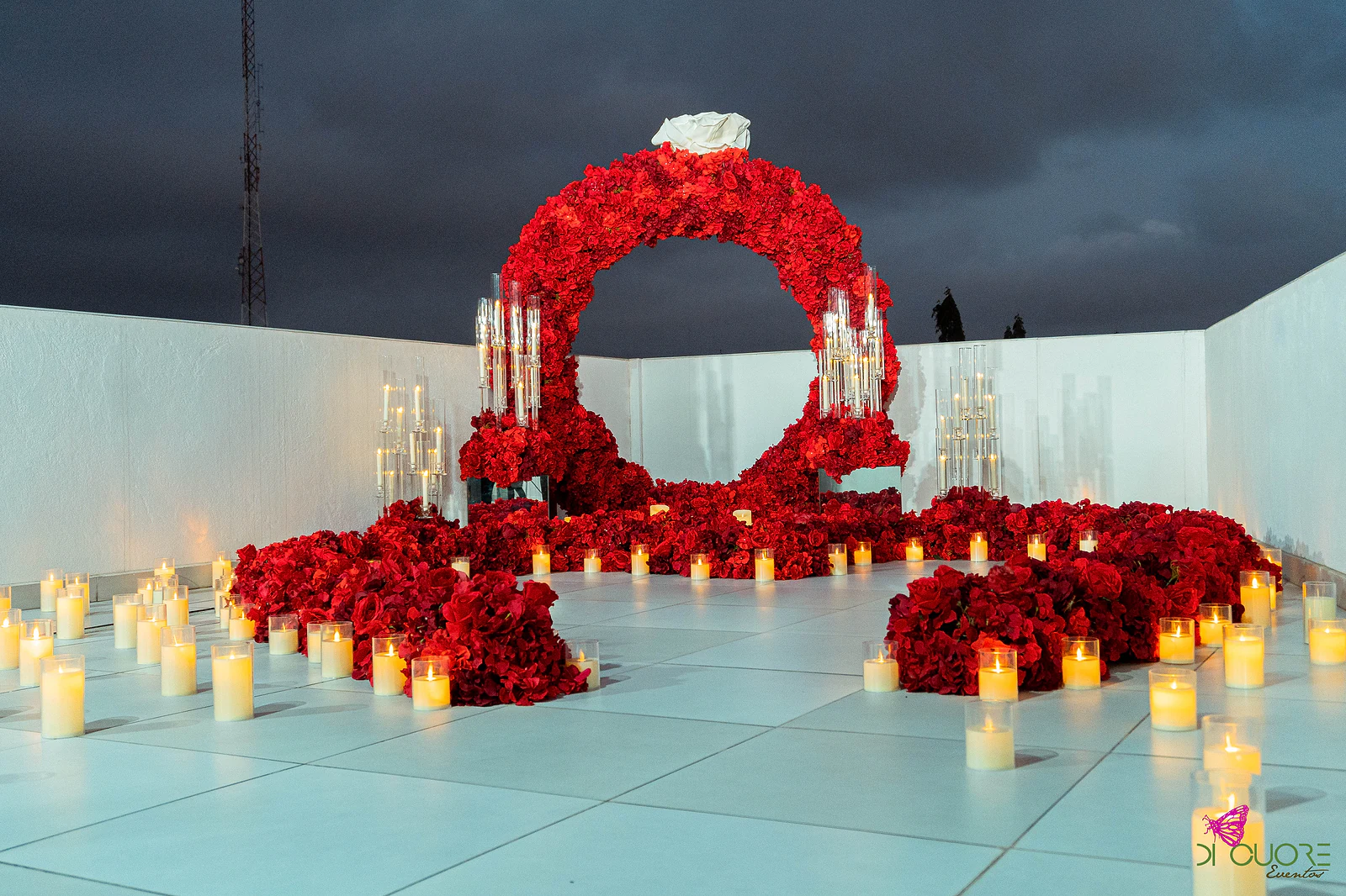 Terraço ao anoitecer com o arco de rosas vermelhas, a rosa branca e dezenas de velas em copos de vidro espalhadas pelo piso claro