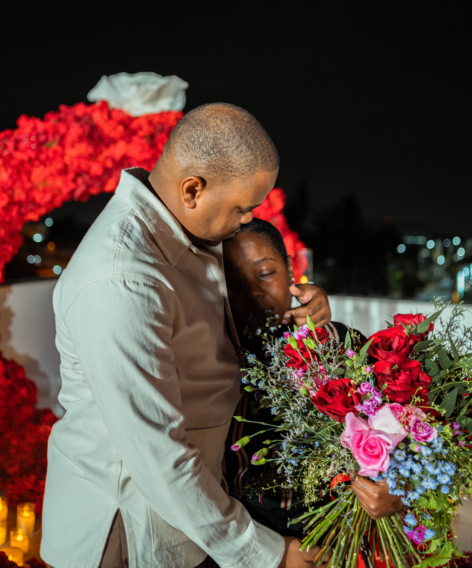 Casal abraçado à noite, ele a pousar o rosto sobre a cabeça dela, com o ramo de rosas e o arco de rosas ao fundo