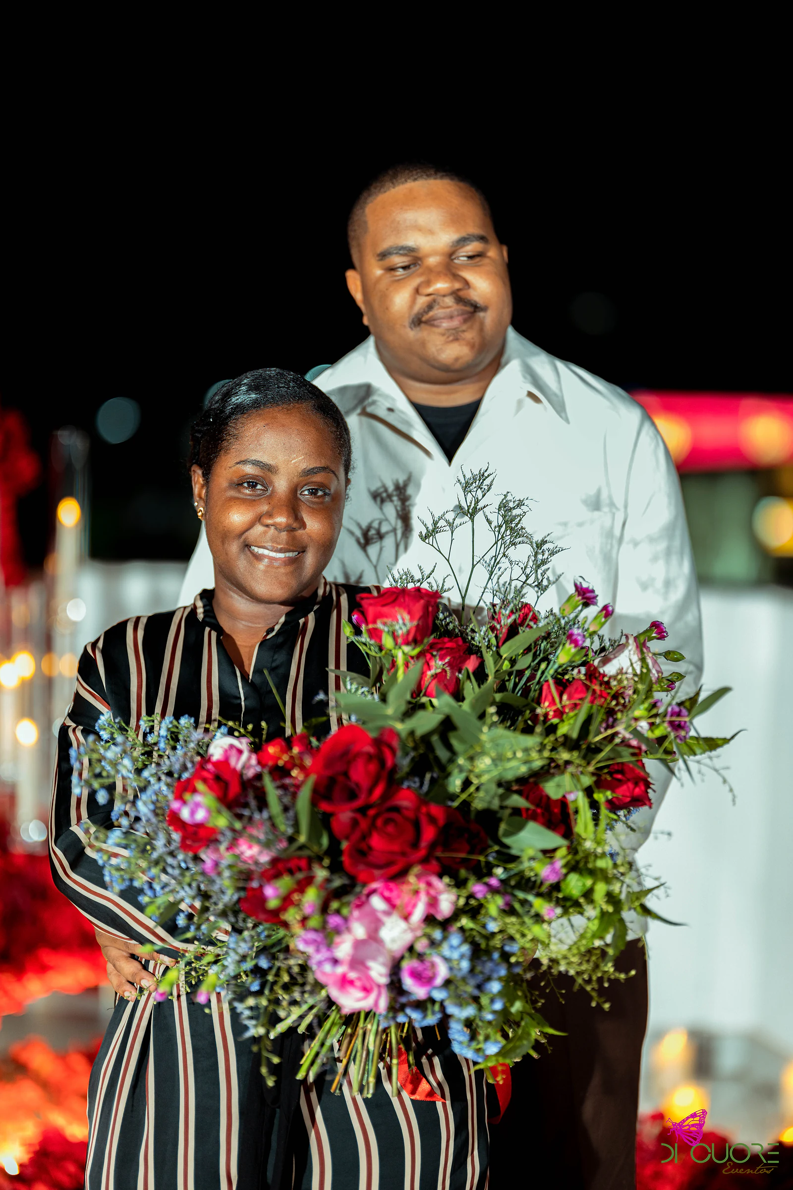 Mulher de vestido às riscas e homem de casaco claro, a sorrir juntos, com um ramo de rosas vermelhas e cor-de-rosa e flores azuis, à noite