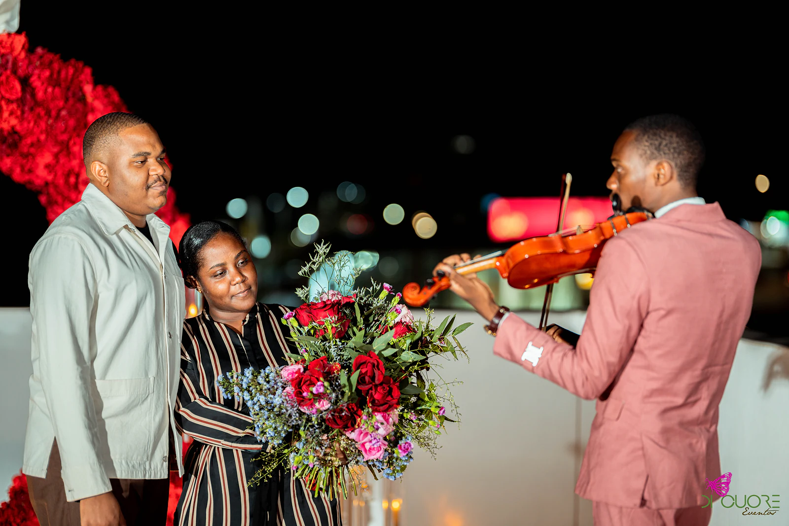 Casal com o ramo de rosas ao lado de um violinista de casaco cor-de-rosa que toca, numa noite de luzes desfocadas