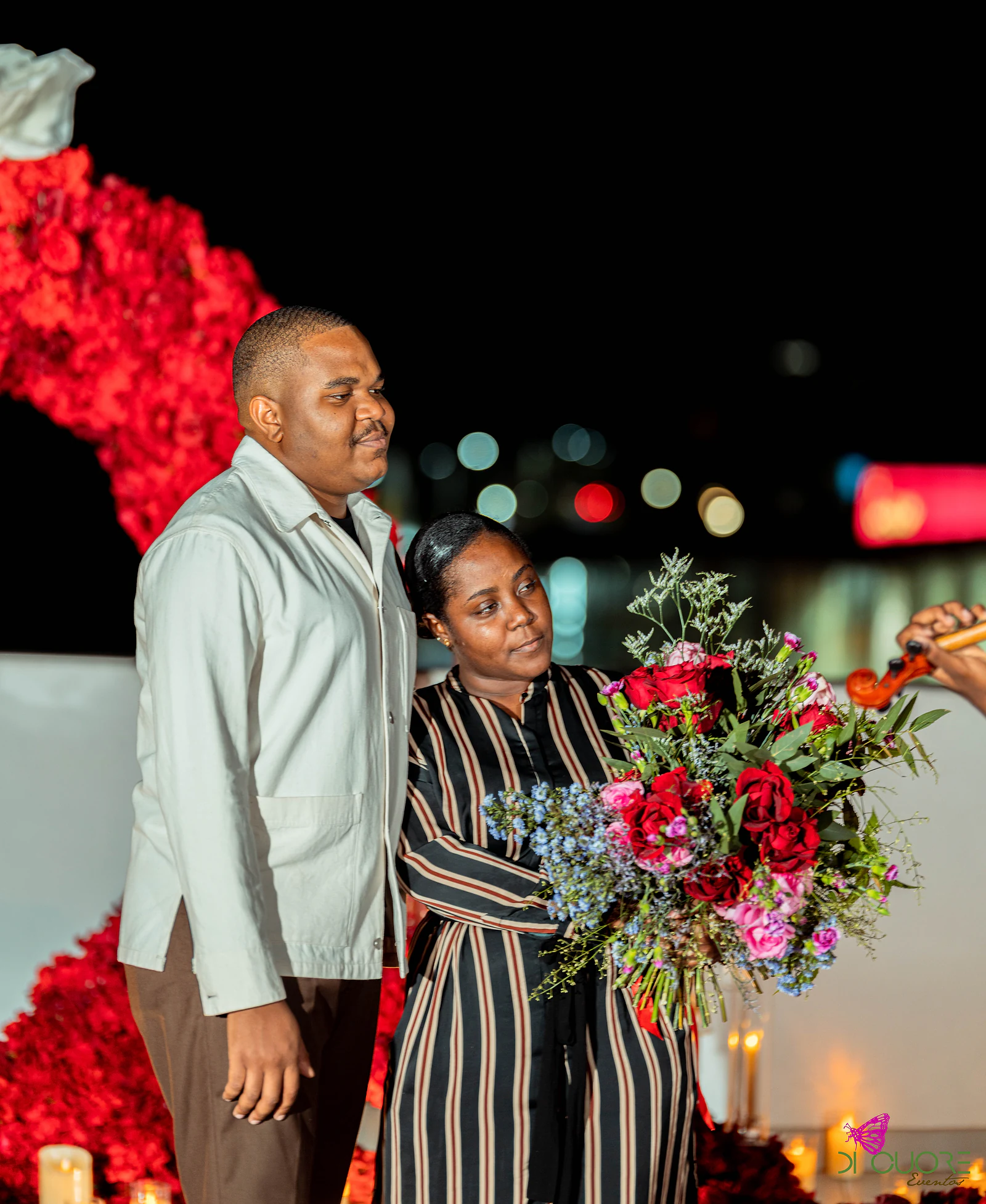 Casal de pé à noite diante do arco de rosas, ele de casaco claro e ela de vestido às riscas com o ramo, e um violino a aparecer à direita