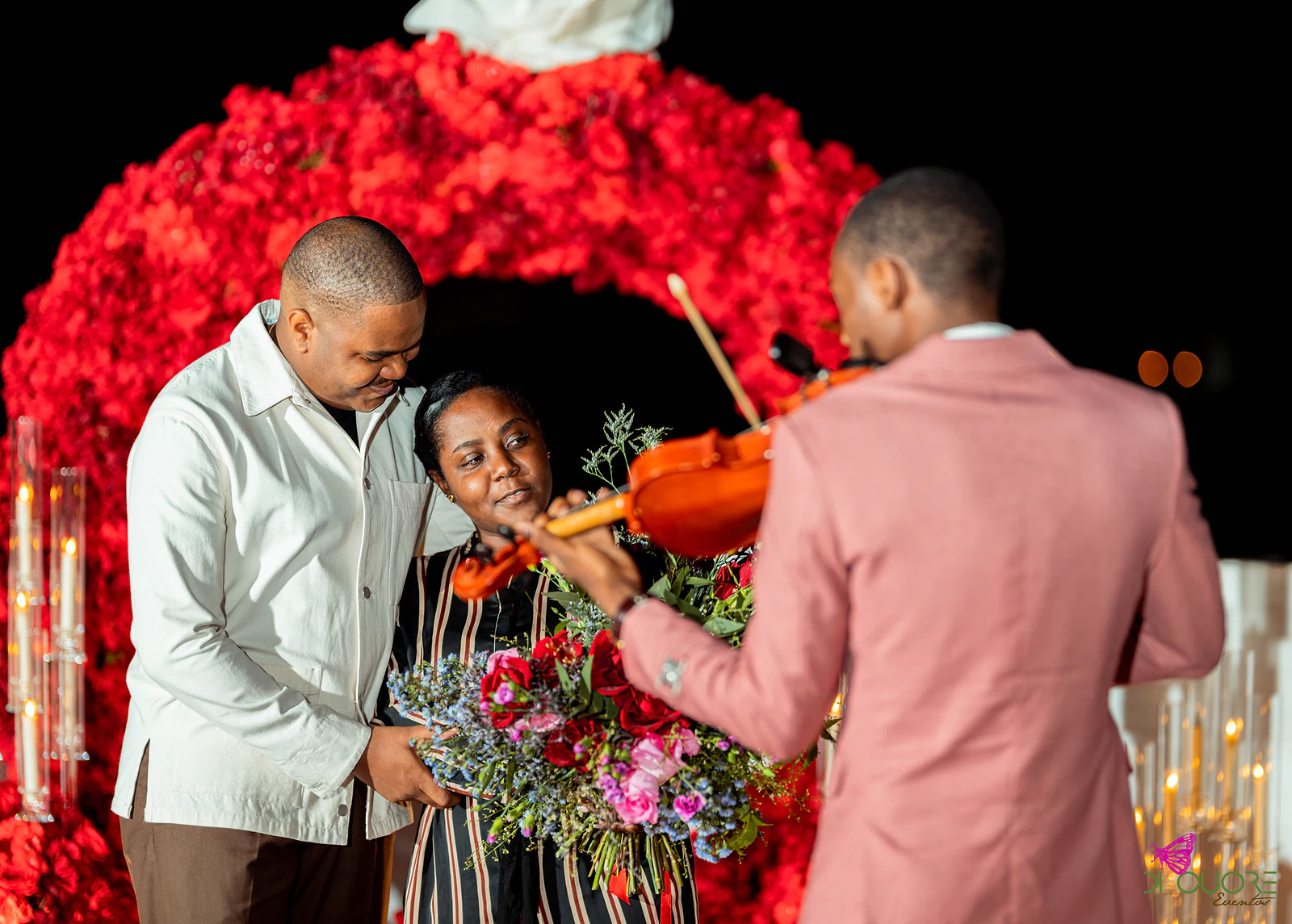 Casal diante do arco de rosas vermelhas, com ela a olhar para ele, e um violinista de casaco cor-de-rosa em primeiro plano