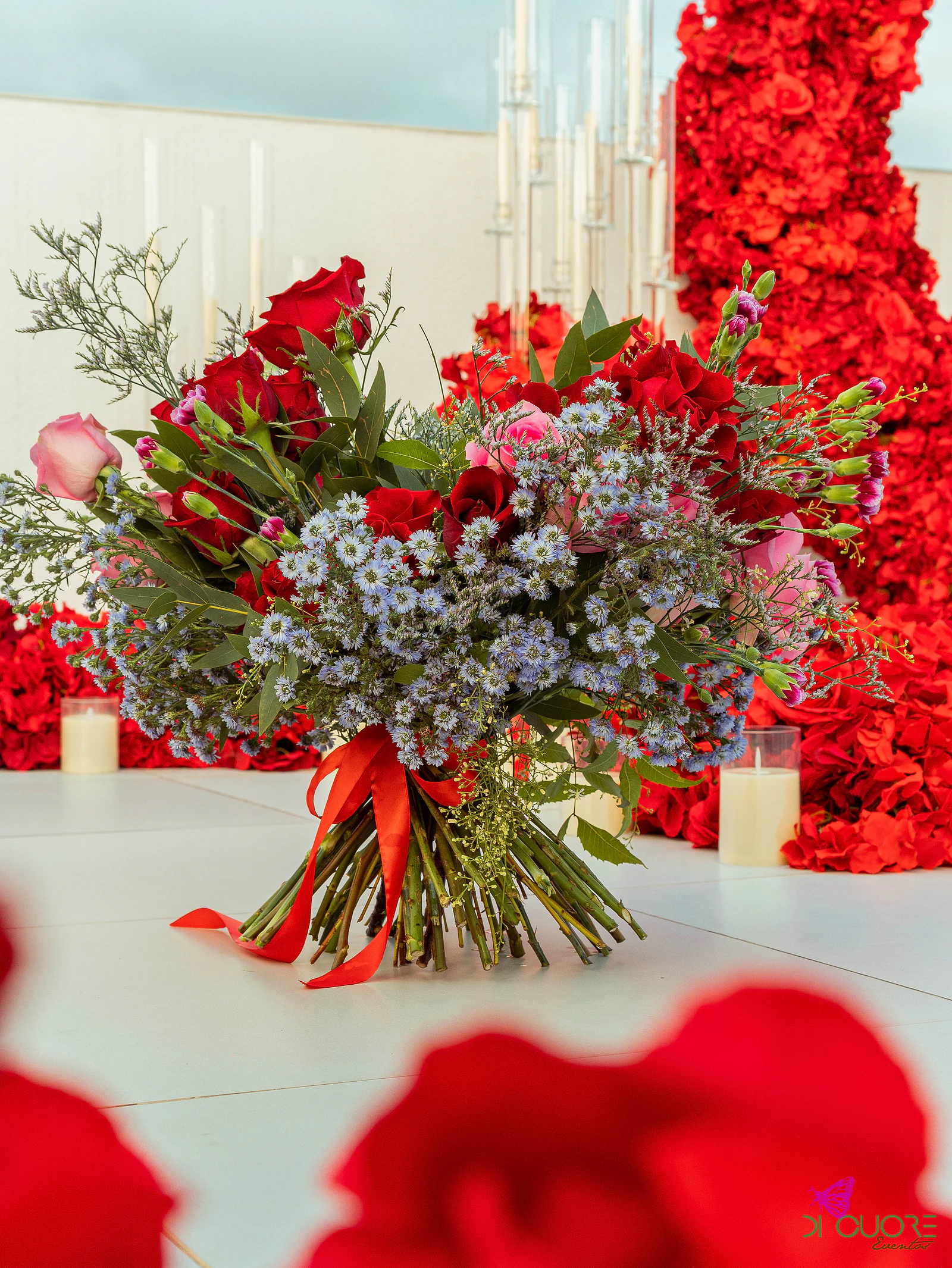 Ramo de rosas vermelhas e cor-de-rosa com flores azuis e fita vermelha, pousado no piso claro, com velas e o arco de flores ao fundo