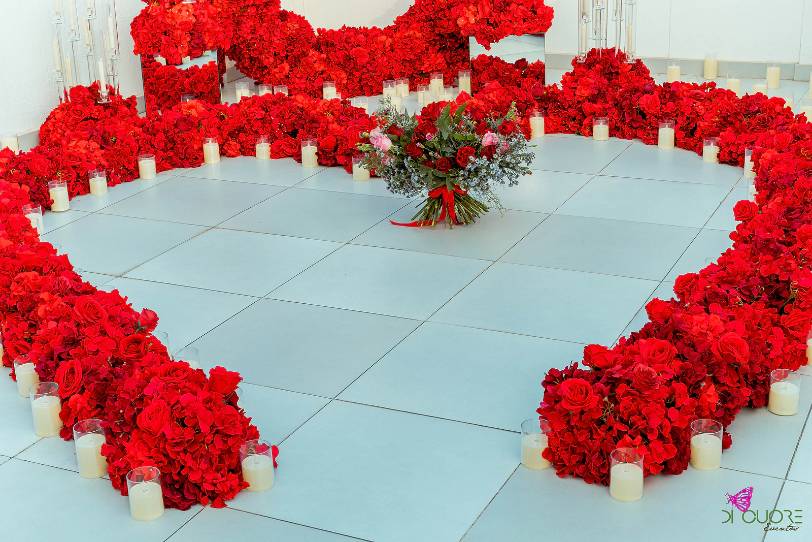 Ramo de rosas e flores azuis pousado no centro de um coração de rosas vermelhas, com velas, sobre o piso claro