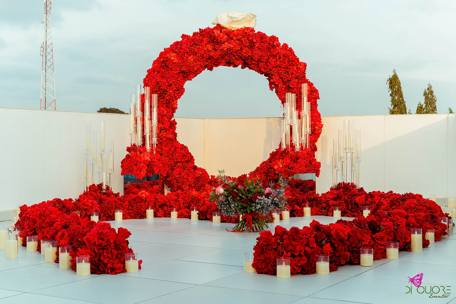 Arco circular de rosas vermelhas rematado por uma rosa branca, com candelabros de vidro, velas e o ramo ao centro do piso claro, sob um céu pálido