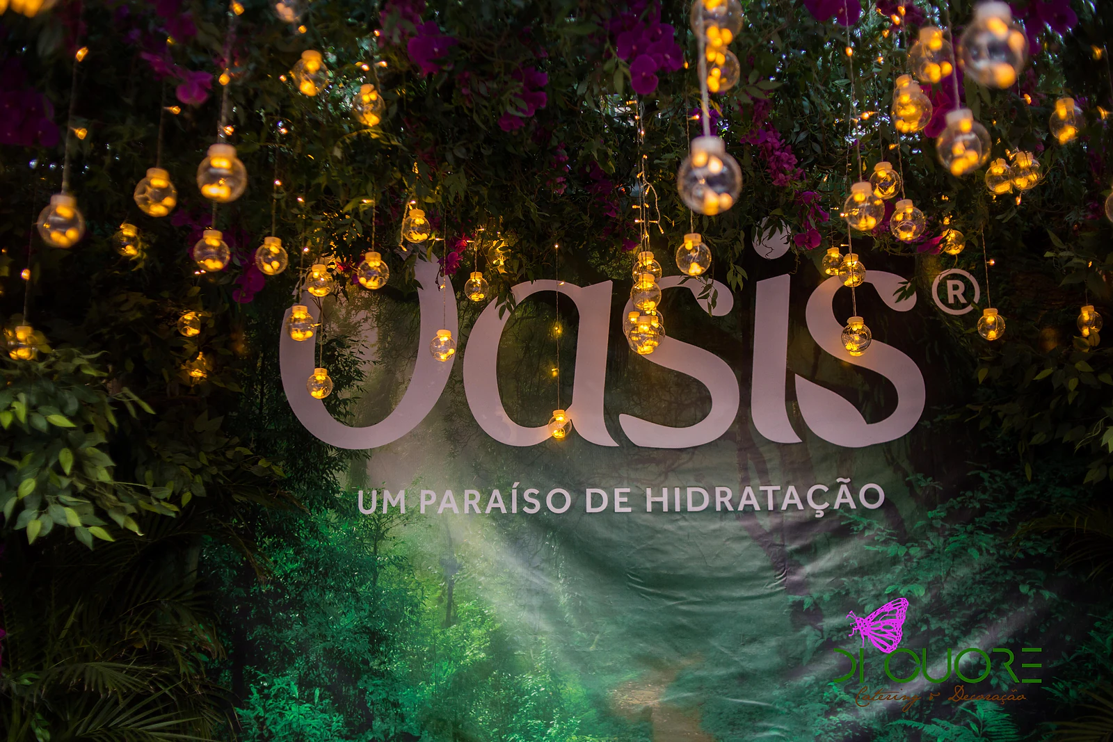 Letreiro Oasis entre folhagem e lâmpadas de vidro acesas, em tons quentes sobre um fundo verde