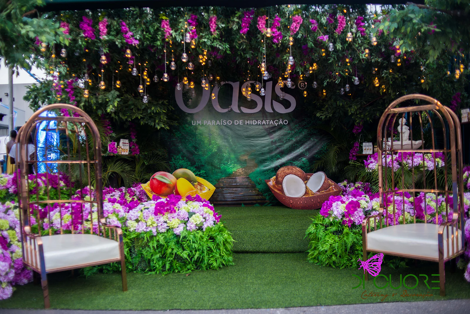 Palco ao ar livre com duas cadeiras em forma de gaiola, relvado, taças de fruta e de coco e um fundo de folhagem, orquídeas e lâmpadas suspensas com o letreiro Oasis