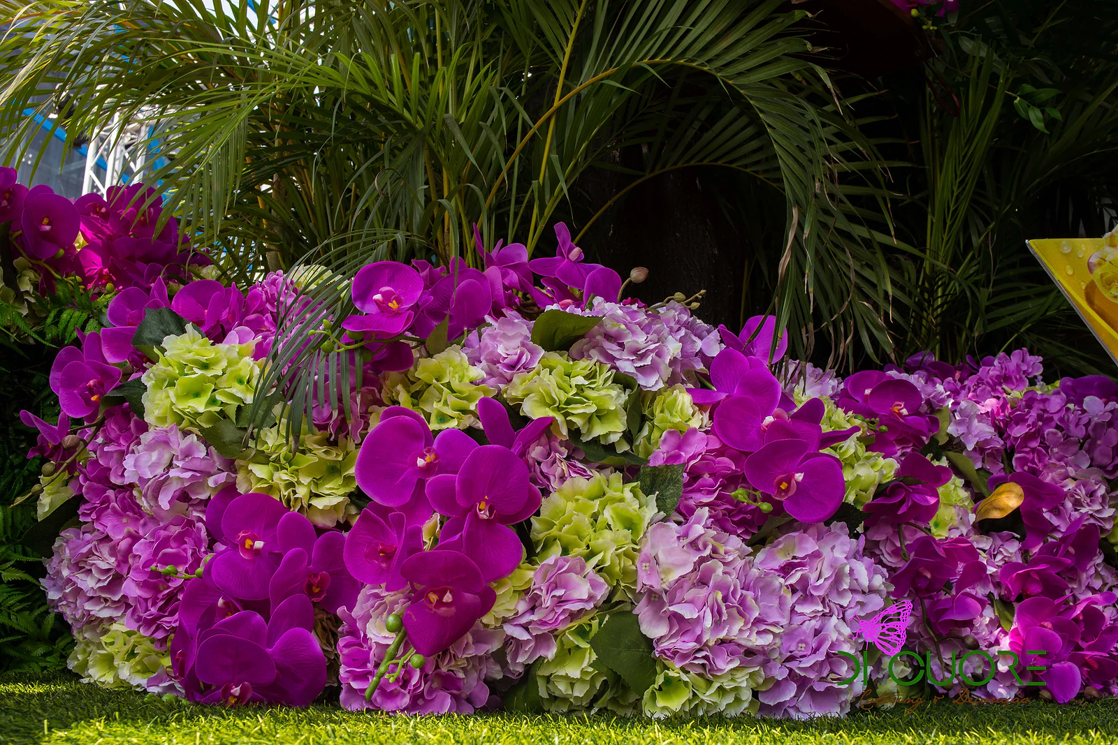 Maciço de orquídeas fúcsia e hortênsias lilás e verde-lima sobre o relvado, com palmeiras por trás