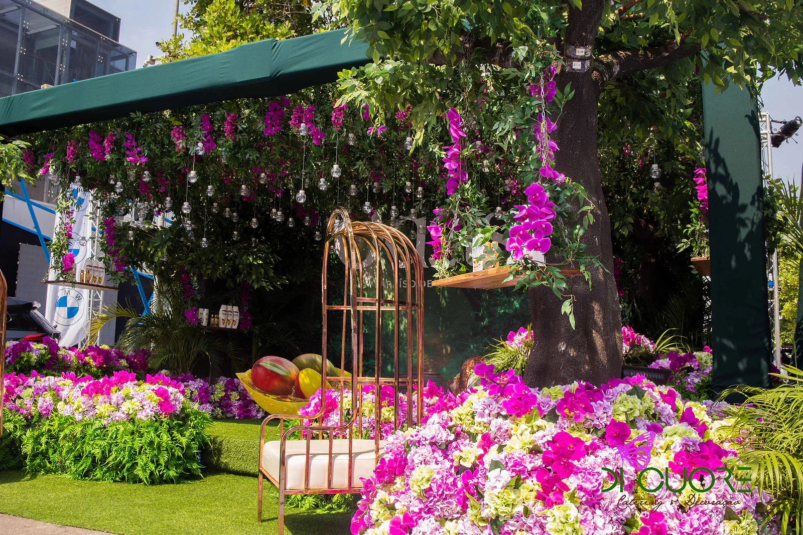 Vista exterior do cenário: tronco de árvore com orquídeas, cadeira em forma de gaiola e maciço de flores cor-de-rosa sob o toldo verde