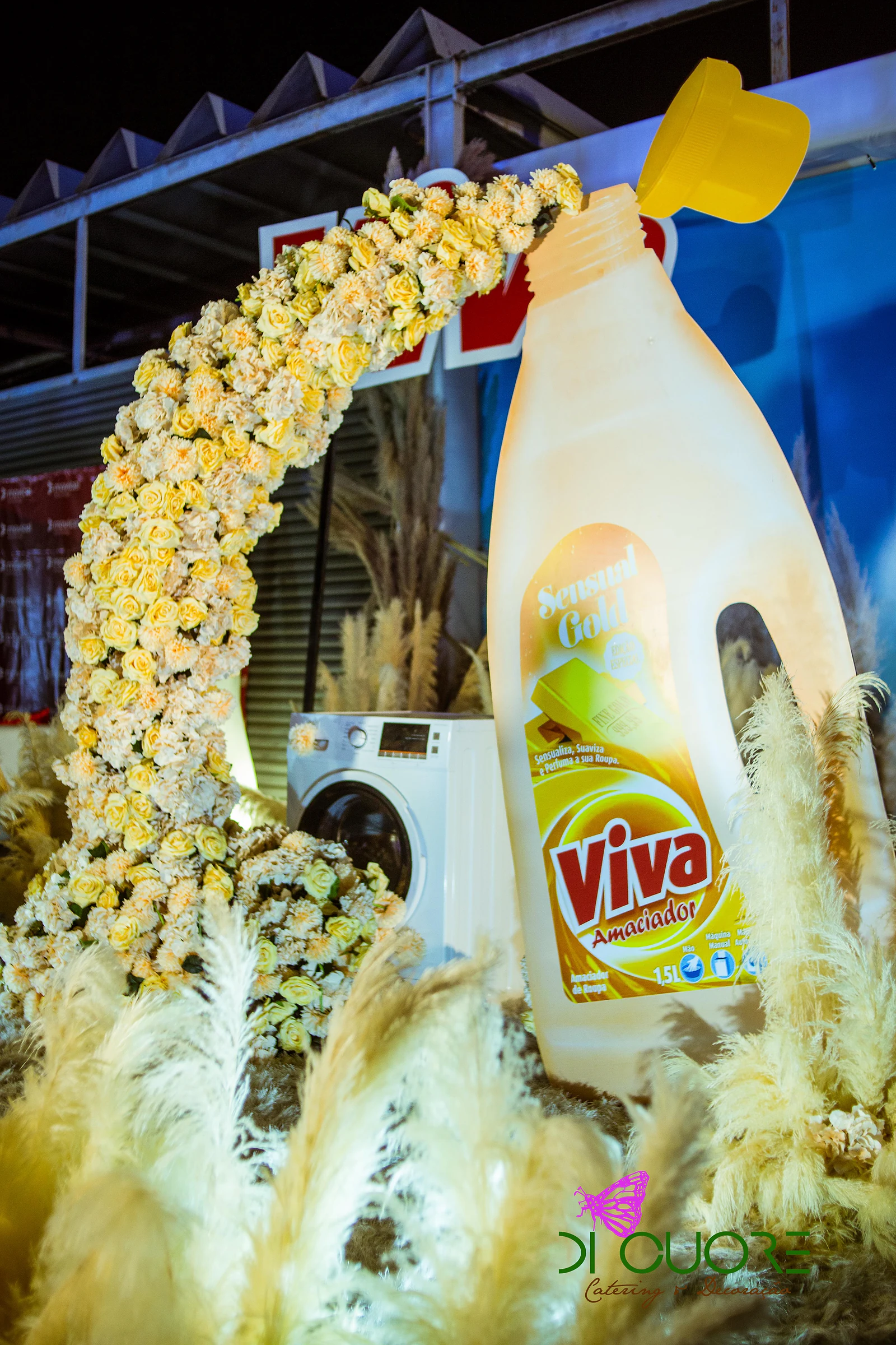 Arco de rosas amarelas a cair da boca de um frasco Viva gigante, com uma máquina de lavar ao fundo e plumas de pampa em primeiro plano