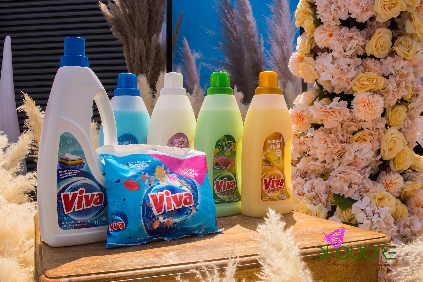 Frascos e saquetas Viva sobre uma mesa de madeira, ao lado de uma coluna de rosas e hortênsias cor de pêssego