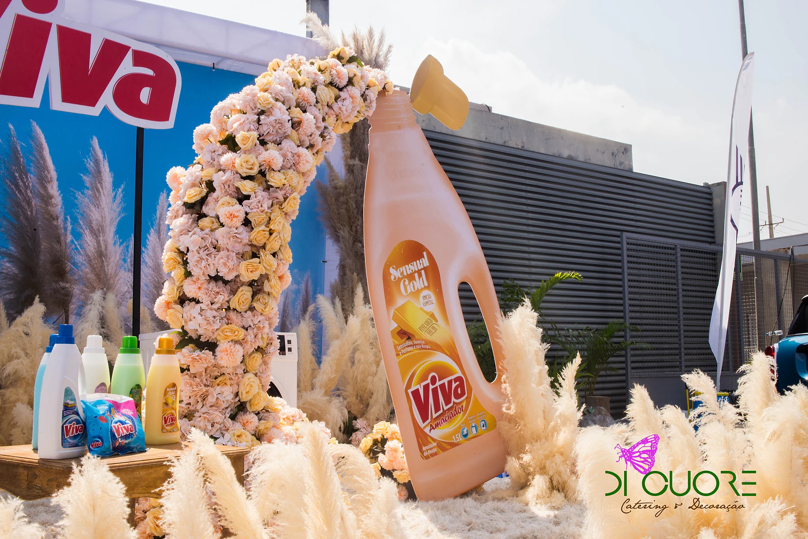 Arco de rosas pêssego e amarelo a verter-se de um frasco Viva gigante, entre plumas de erva-das-pampas e um fundo de céu azul