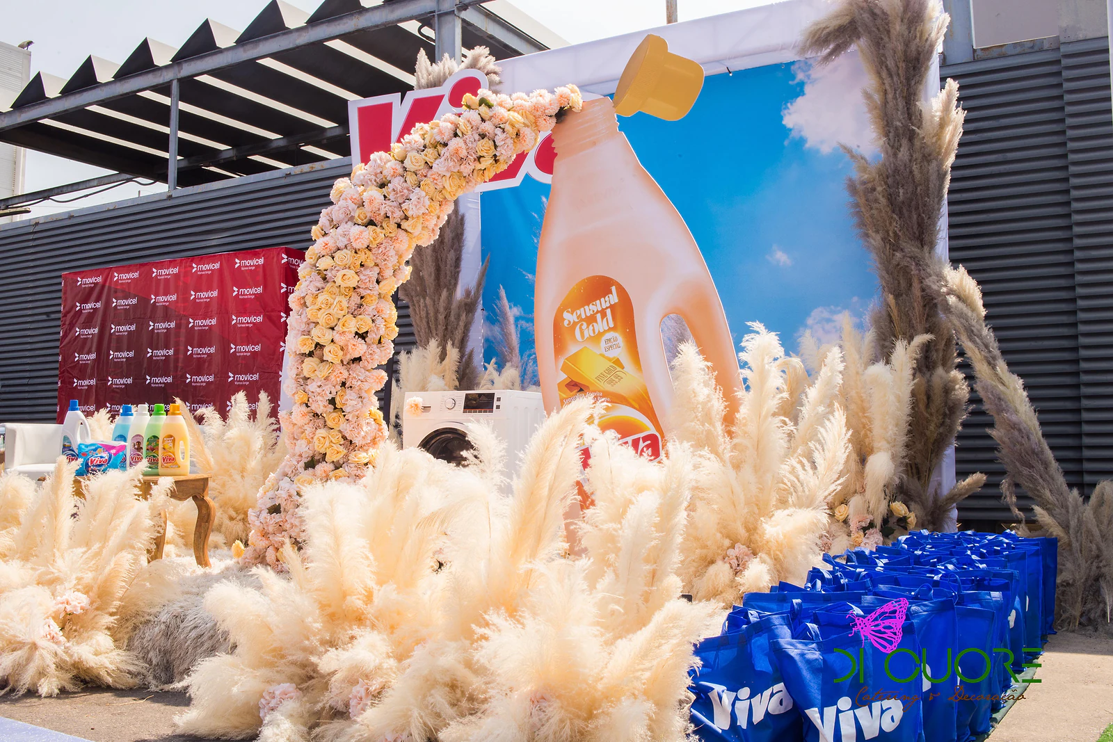 Arch of peach and yellow roses pouring from a giant bottle, amid plumes of pampas grass, with blue bags in the foreground