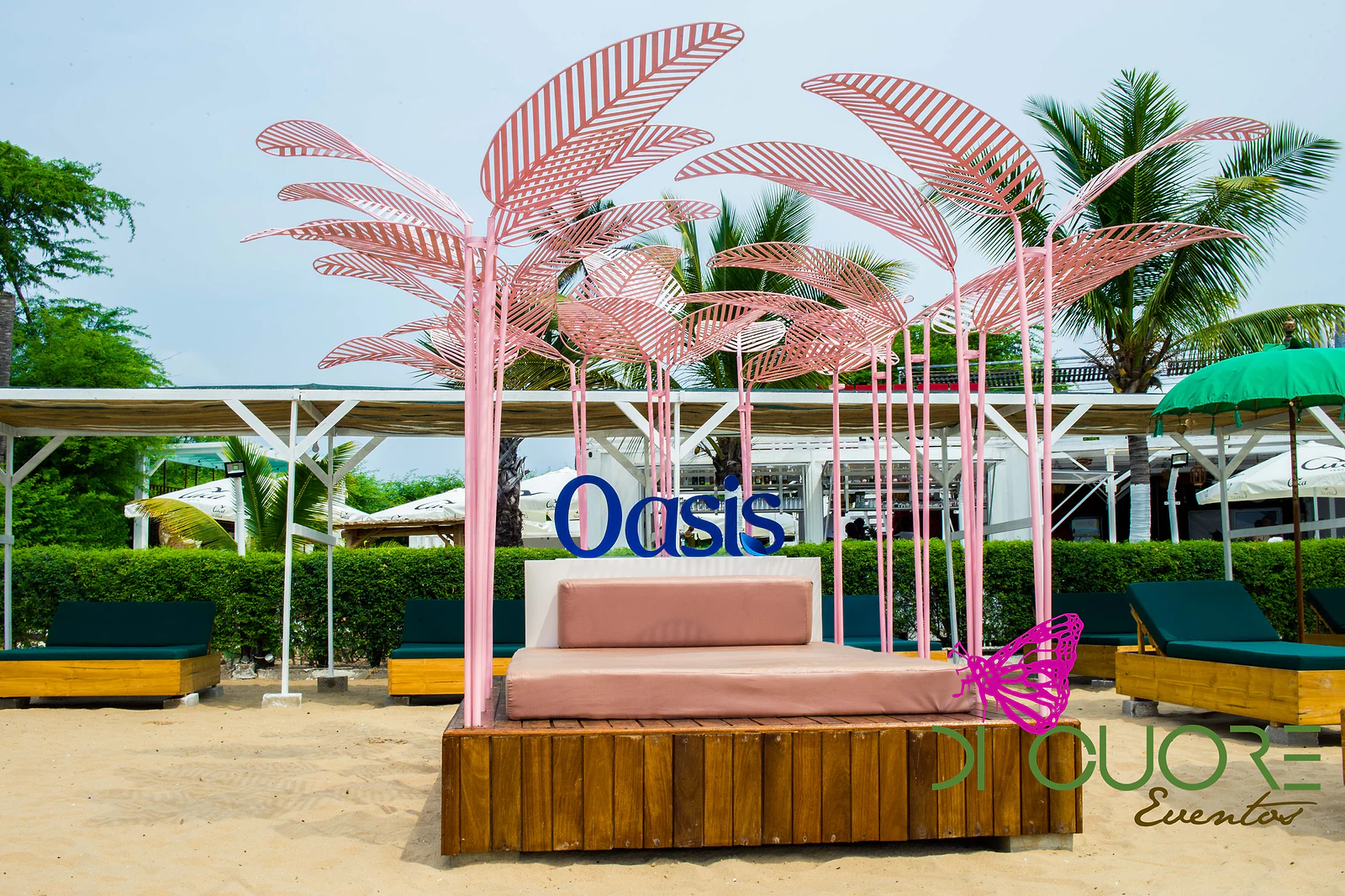 Cama de deck com almofadas rosa-velho sob um dossel de folhas cor-de-rosa e o nome da marca em azul, entre espreguiçadeiras verdes e palmeiras