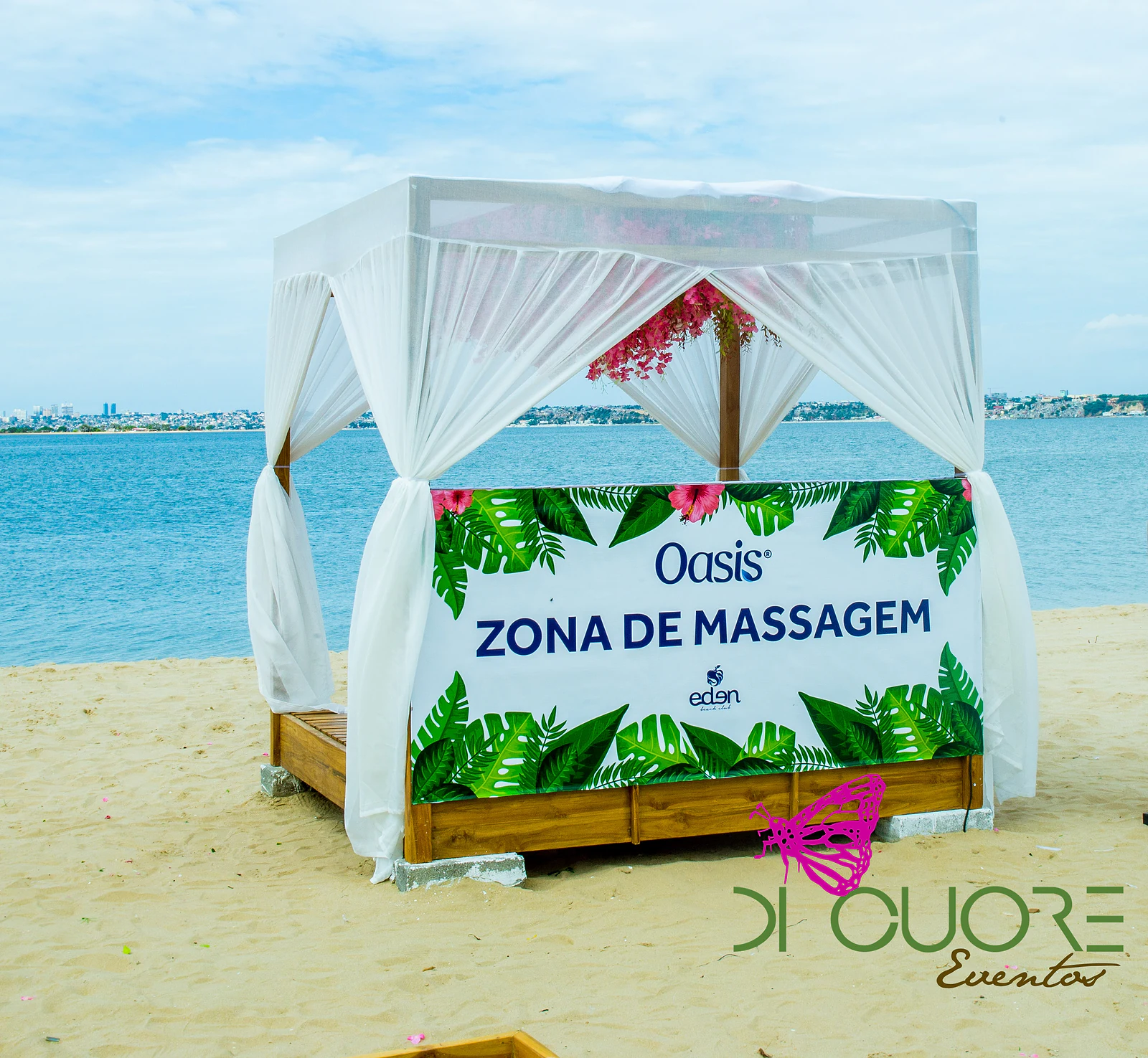 Cabana branca de cortinados leves na areia, com o letreiro Zona de Massagem emoldurado por folhas tropicais e o mar ao fundo
