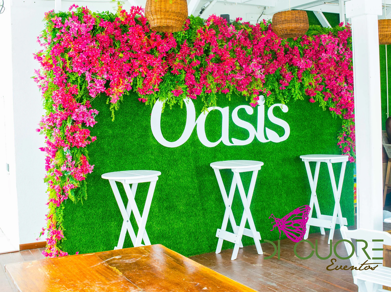 Parede de relva verde com o nome da marca em branco, coroada de flores cor-de-rosa, com bancos altos brancos à frente