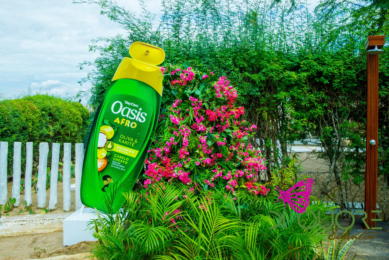 Frasco gigante da marca Oasis entre uma parede de folhagem verde e flores cor-de-rosa e vermelhas, com fetos em primeiro plano