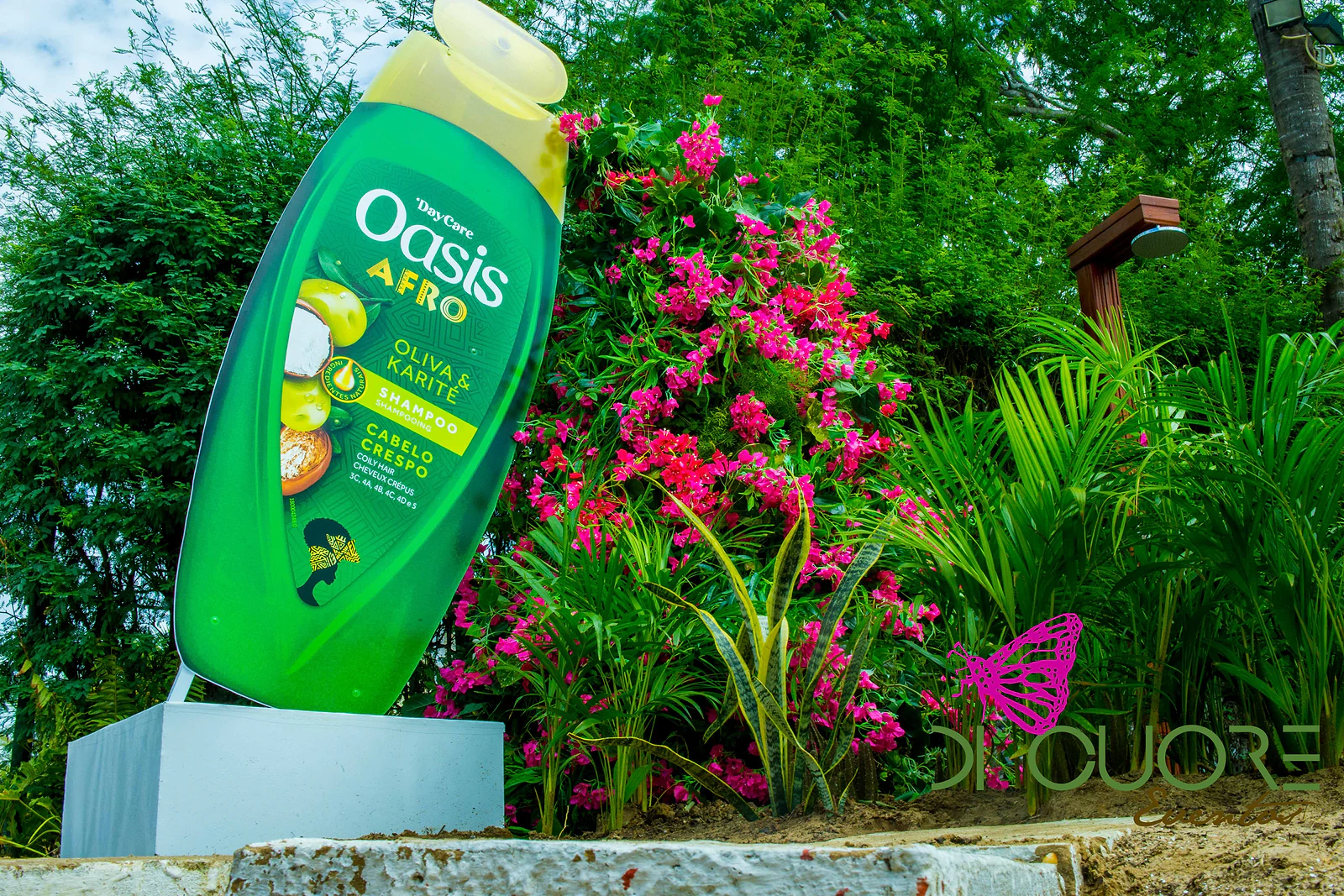 Frasco gigante do produto da marca entre palmeiras e arbustos de flores cor-de-rosa