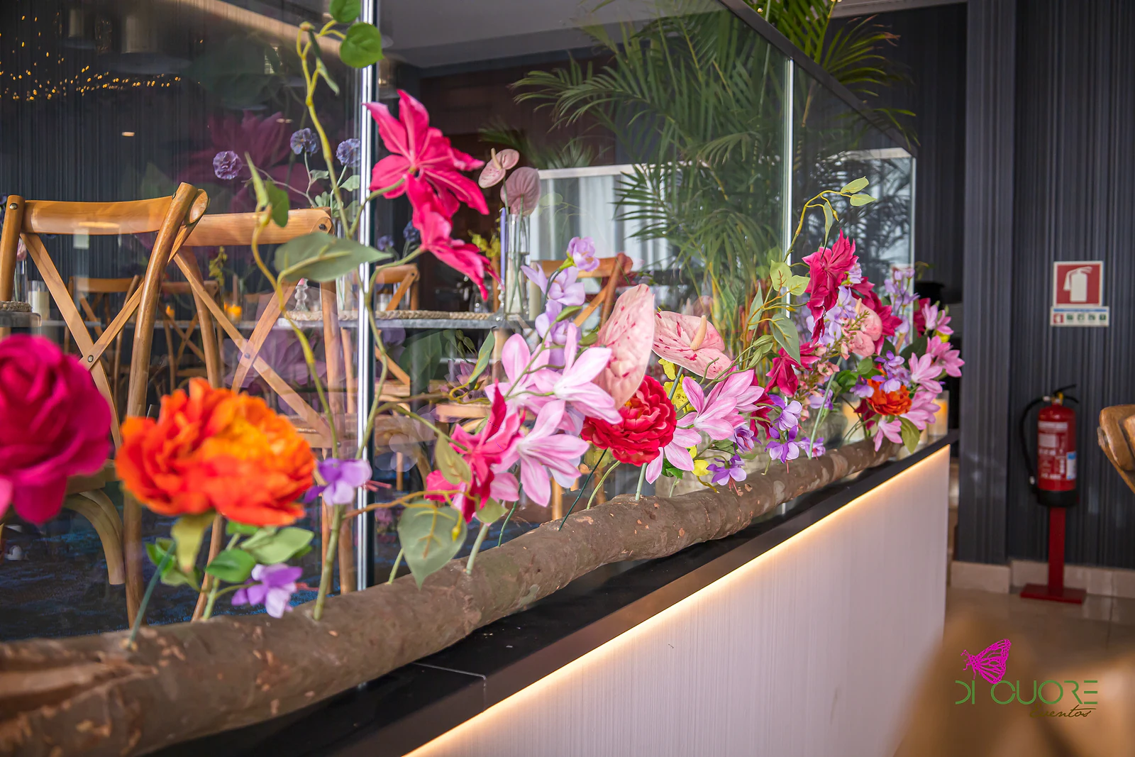 Ramos de flores cor-de-rosa, laranja e roxas num tronco sobre o balcão de vidro, com luz quente por baixo