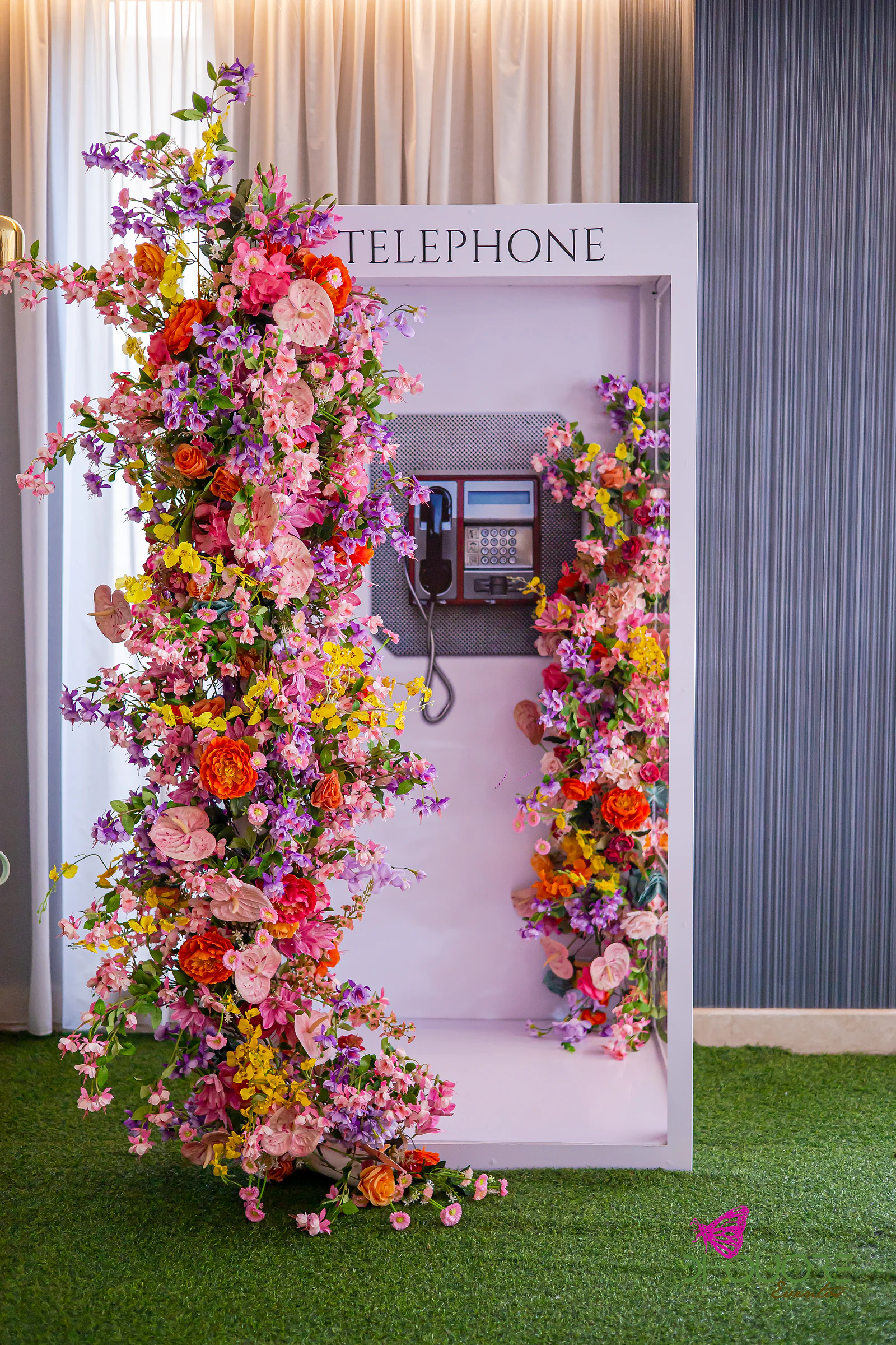 Cabine telefónica branca com uma cascata de flores cor-de-rosa, laranja, roxas e amarelas e antúrios, sobre relva sintética