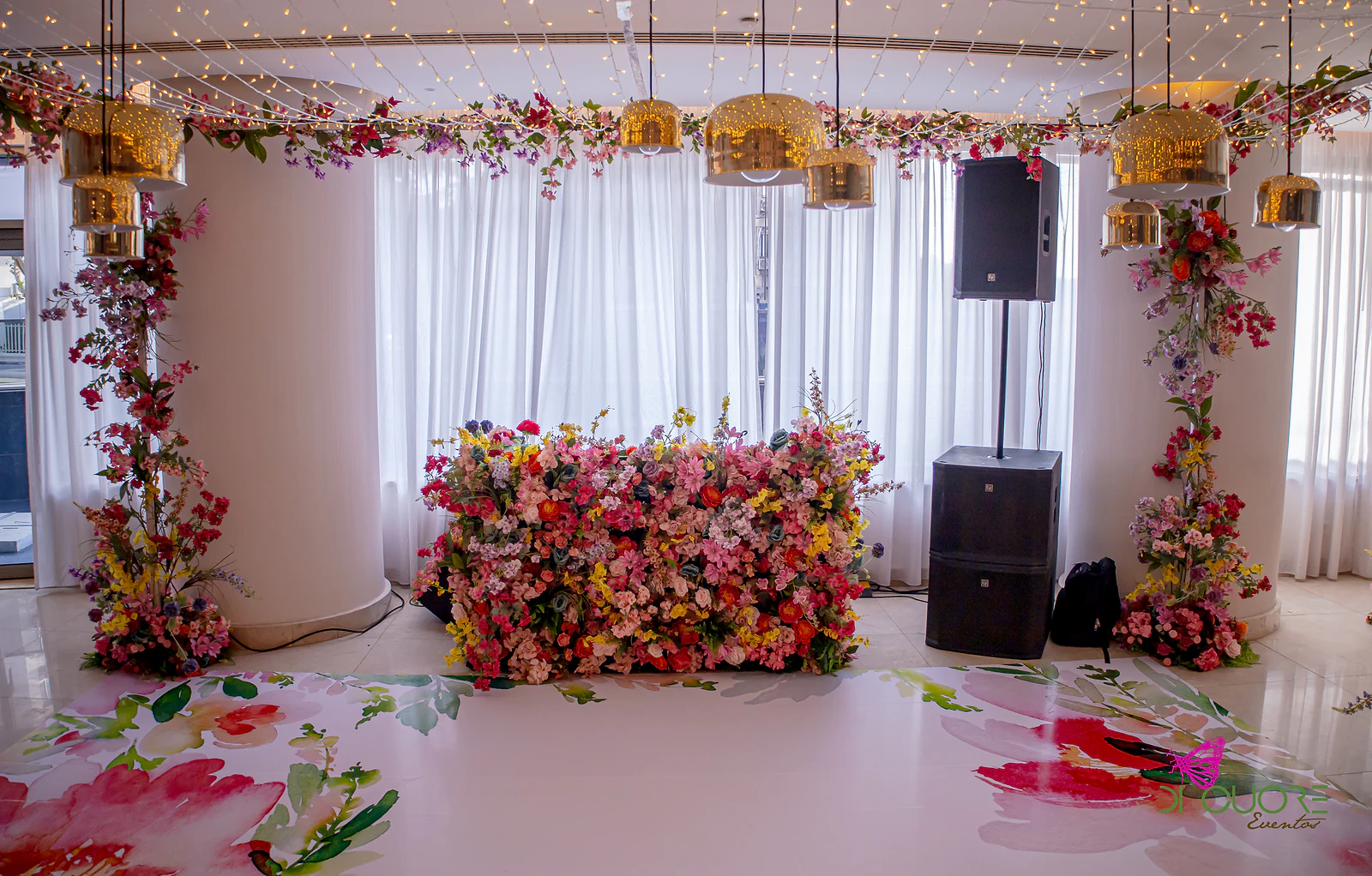 Muro de flores de cor entre duas colunas brancas, com candeeiros dourados, cortinados brancos e colunas de flores de cada lado