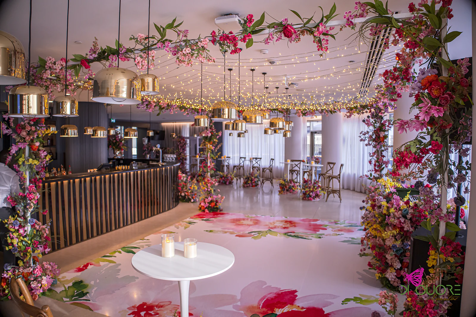 Balcão curvo de ripas de madeira sob candeeiros dourados e um tecto de luzes, com uma mesa branca com velas na pista de flores