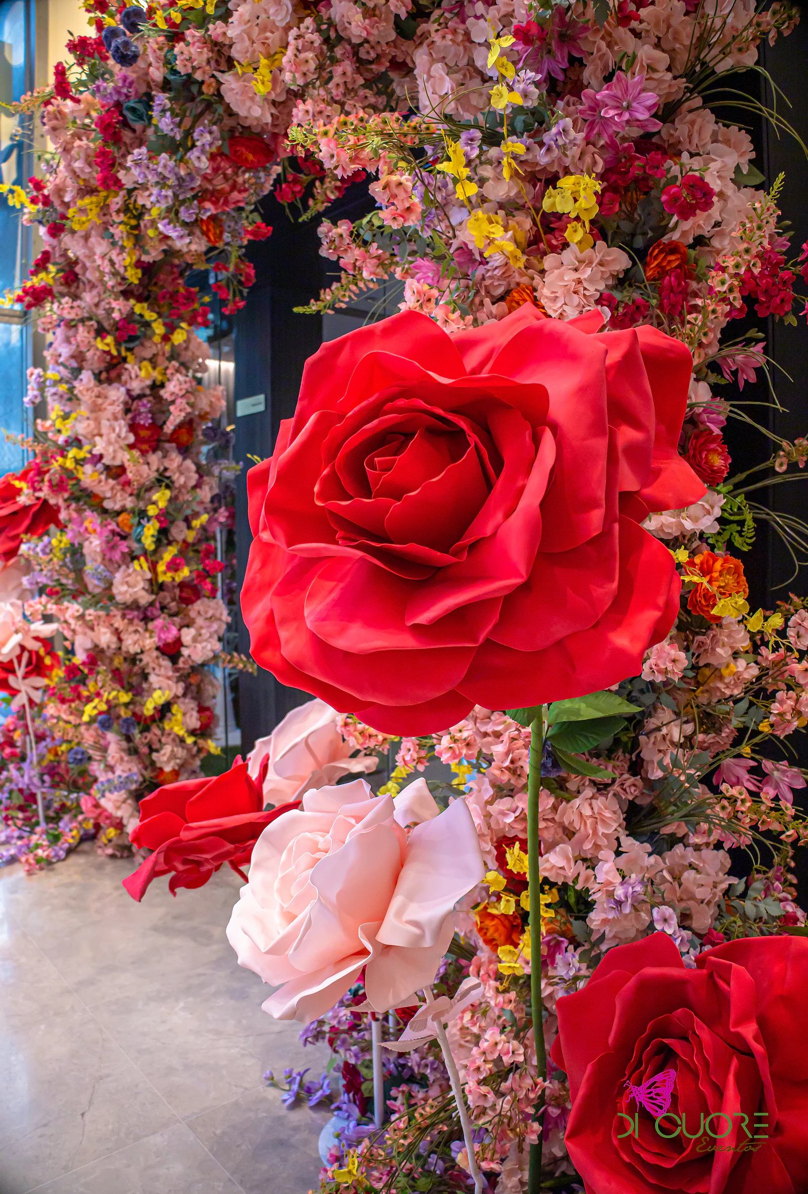 Rosa gigante vermelha e rosas gigantes cor-de-rosa em primeiro plano, diante de uma parede de flores de cor