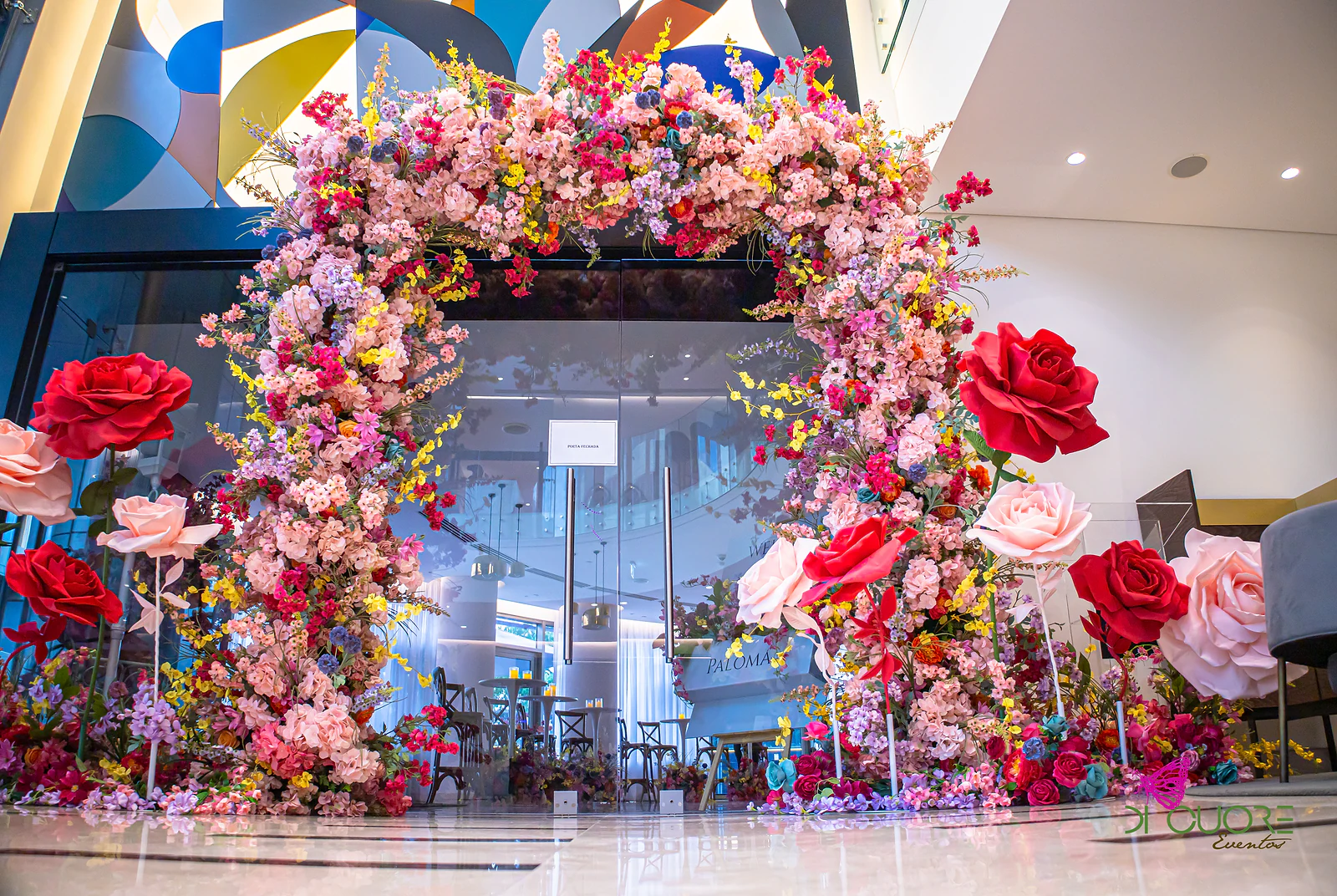 Arco de flores cor-de-rosa, vermelhas, roxas e amarelas à entrada, com rosas gigantes dos lados e uma placa ao centro