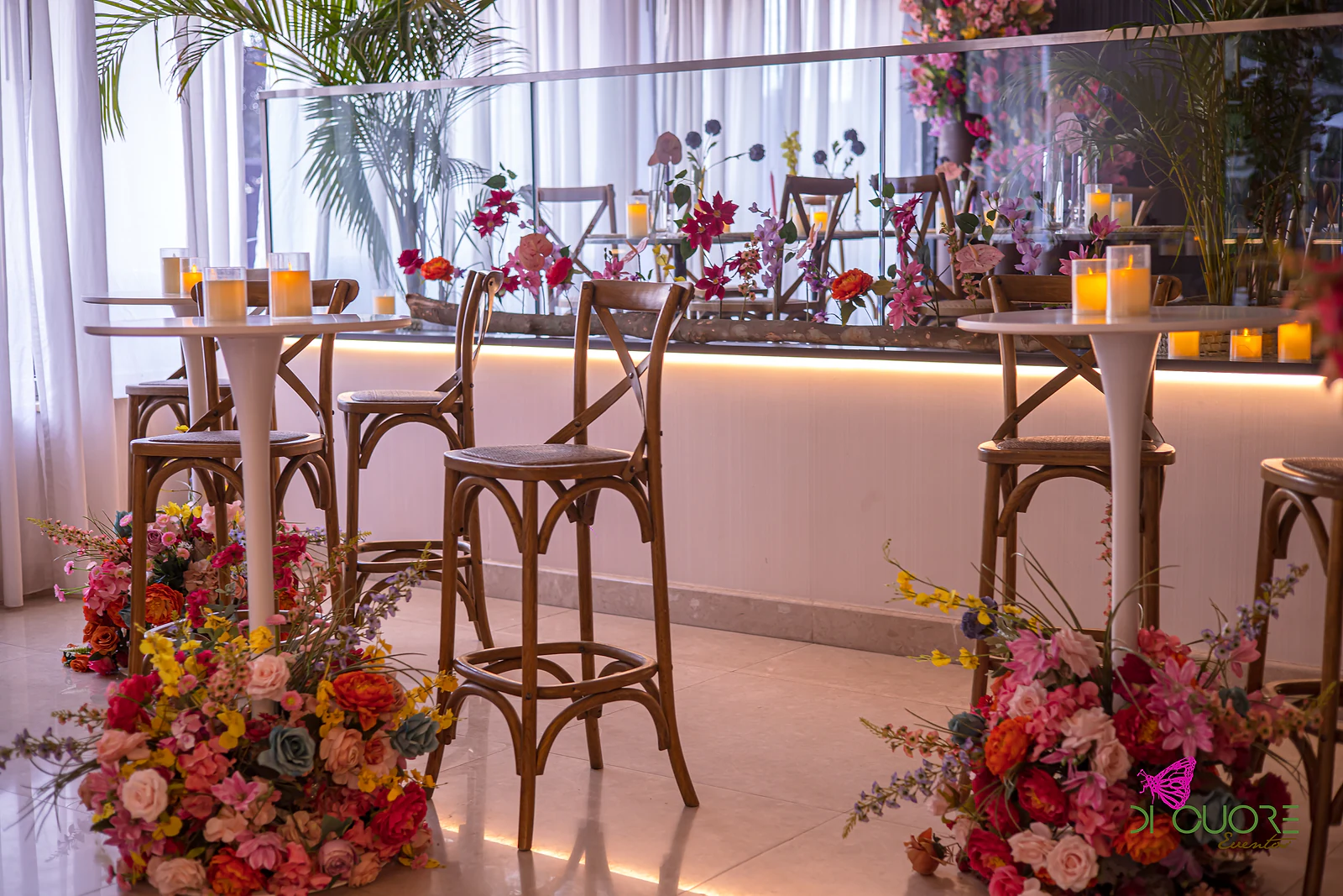 Bancos altos de madeira e mesas brancas com velas, com ramos de flores cor-de-rosa e amarelas ao pé e um balcão com luz por baixo