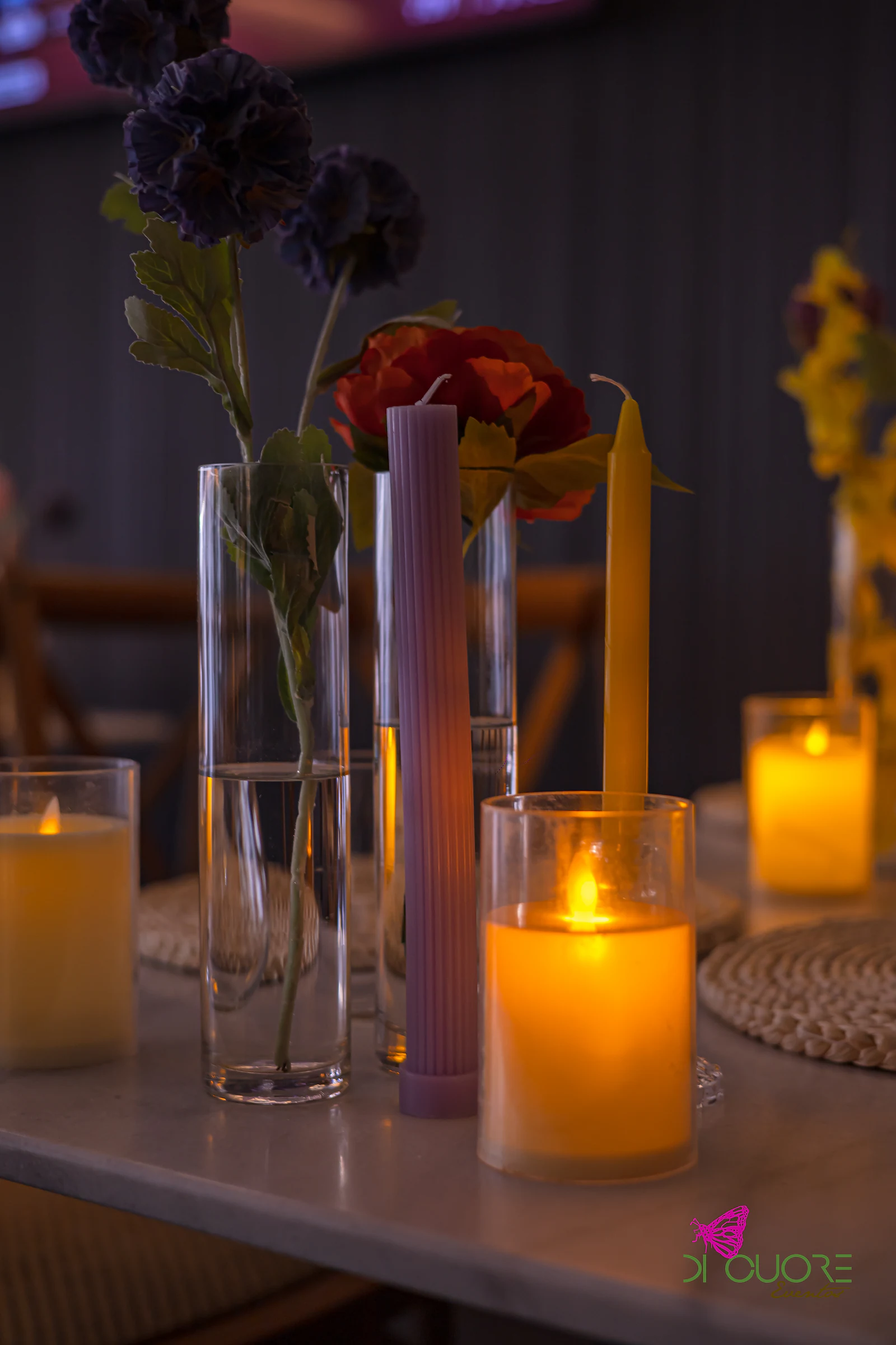 Velas em copos de vidro, jarras com flores roxas e laranja e uma vela lilás, sobre uma mesa de tampo claro