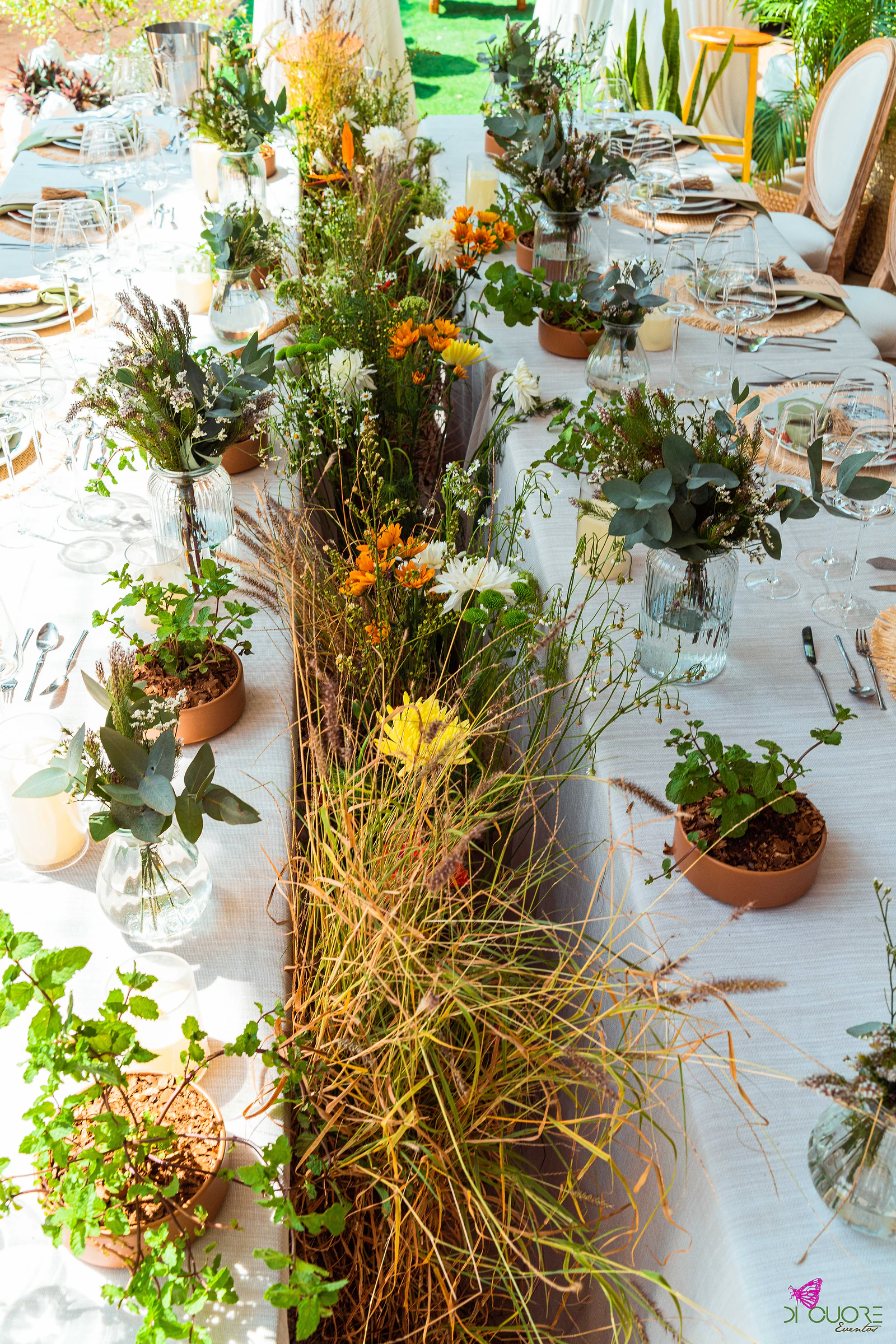 Mesa vista de cima, com uma faixa de capim e flores do campo ao centro, vasos de barro com plantas, ramos de eucalipto e velas