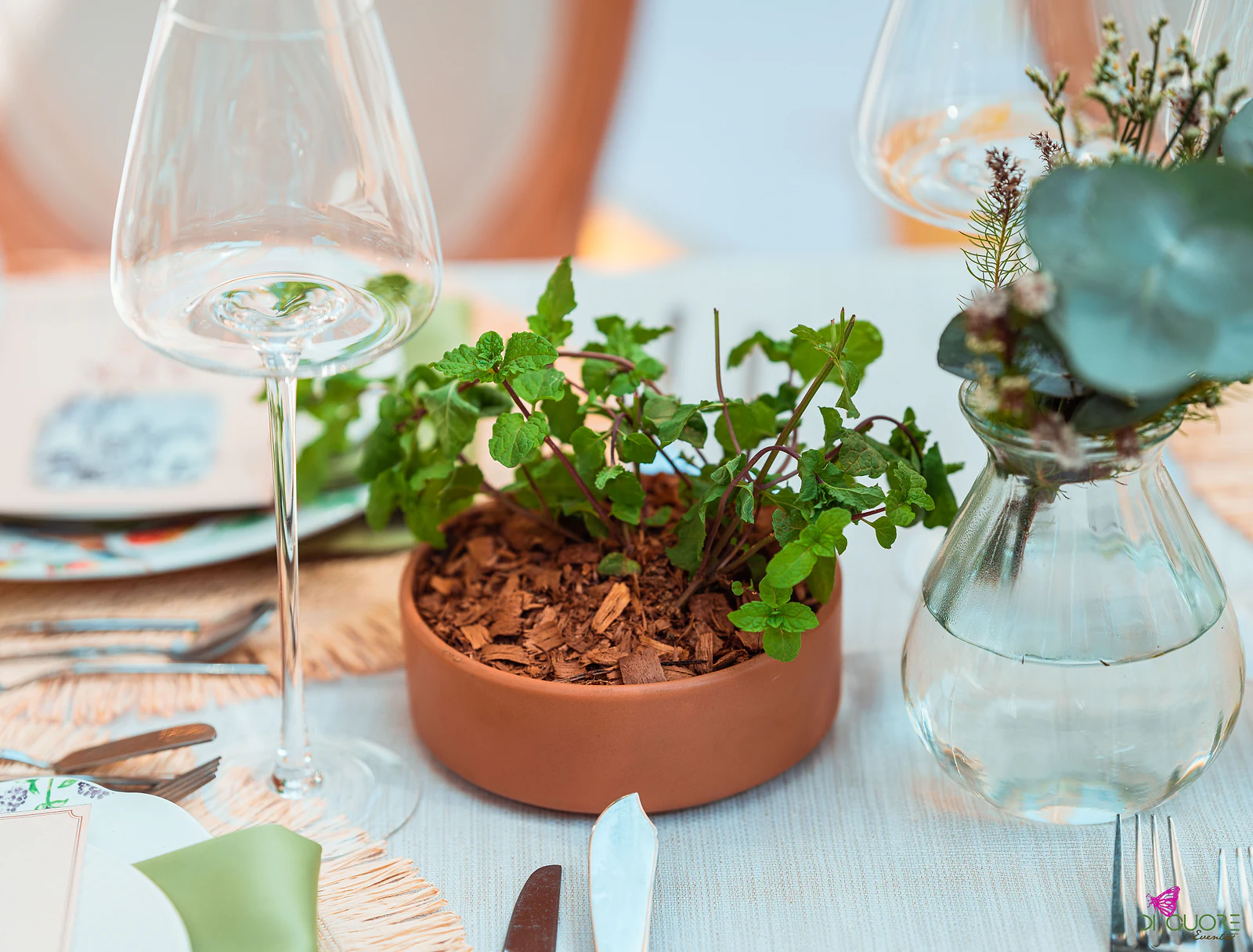 Vaso de barro baixo com plantas e jarra de vidro com eucalipto, junto de copos e talheres