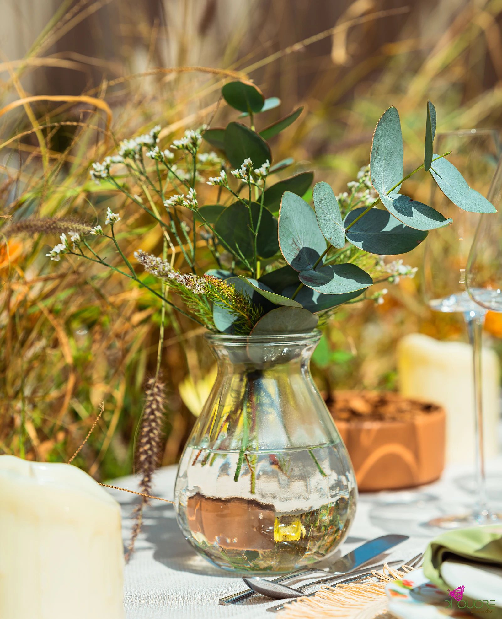 Jarra de vidro com eucalipto e pequenas flores brancas, entre velas e capim seco, com um vaso de barro ao fundo