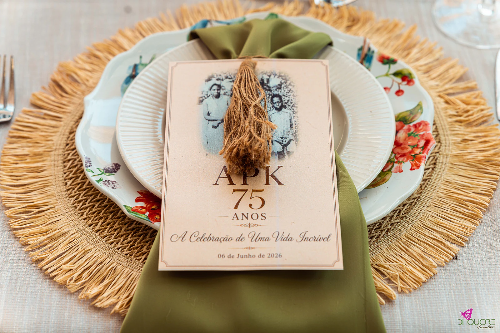 Prato de porcelana com flores pintadas sobre individual de ráfia, guardanapo verde-oliva e ementa com o monograma APK 75 anos