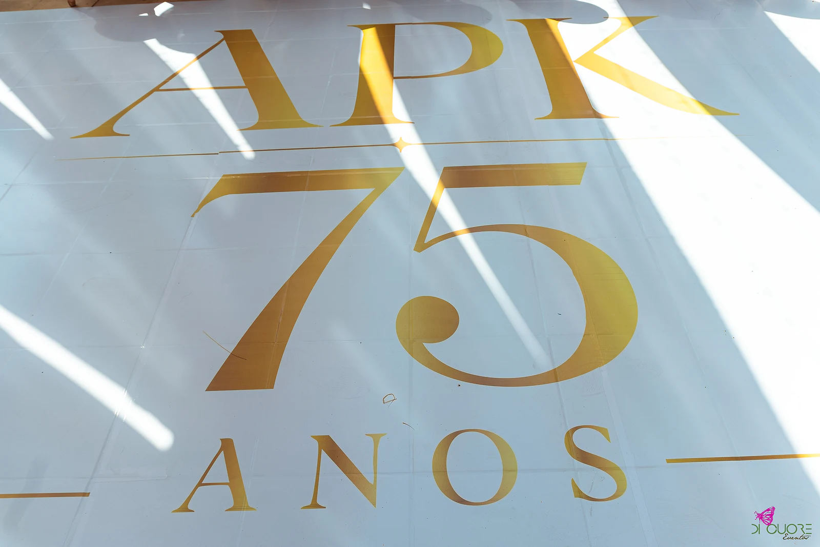 Monograma dourado APK 75 anos sobre fundo branco, com a sombra dos cortinados