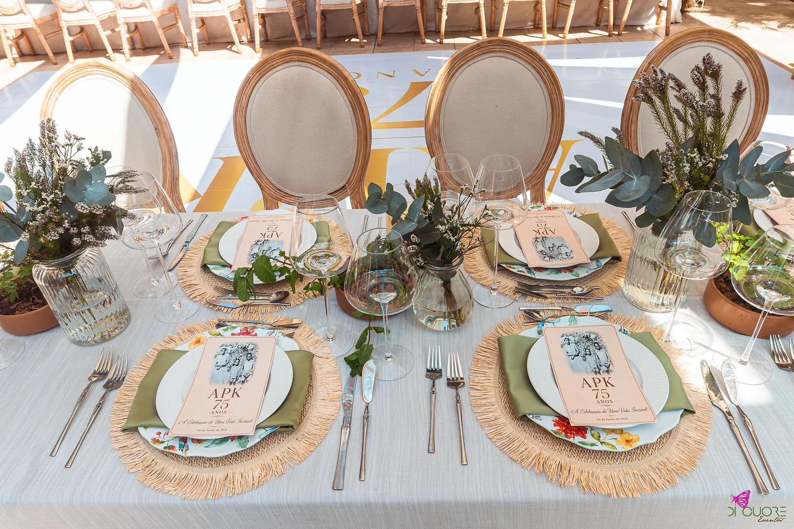Três lugares postos com pratos de porcelana floral, individuais de ráfia, guardanapos verde-oliva e ementas APK 75 anos, com jarras de eucalipto e cadeiras de encosto oval ao fundo