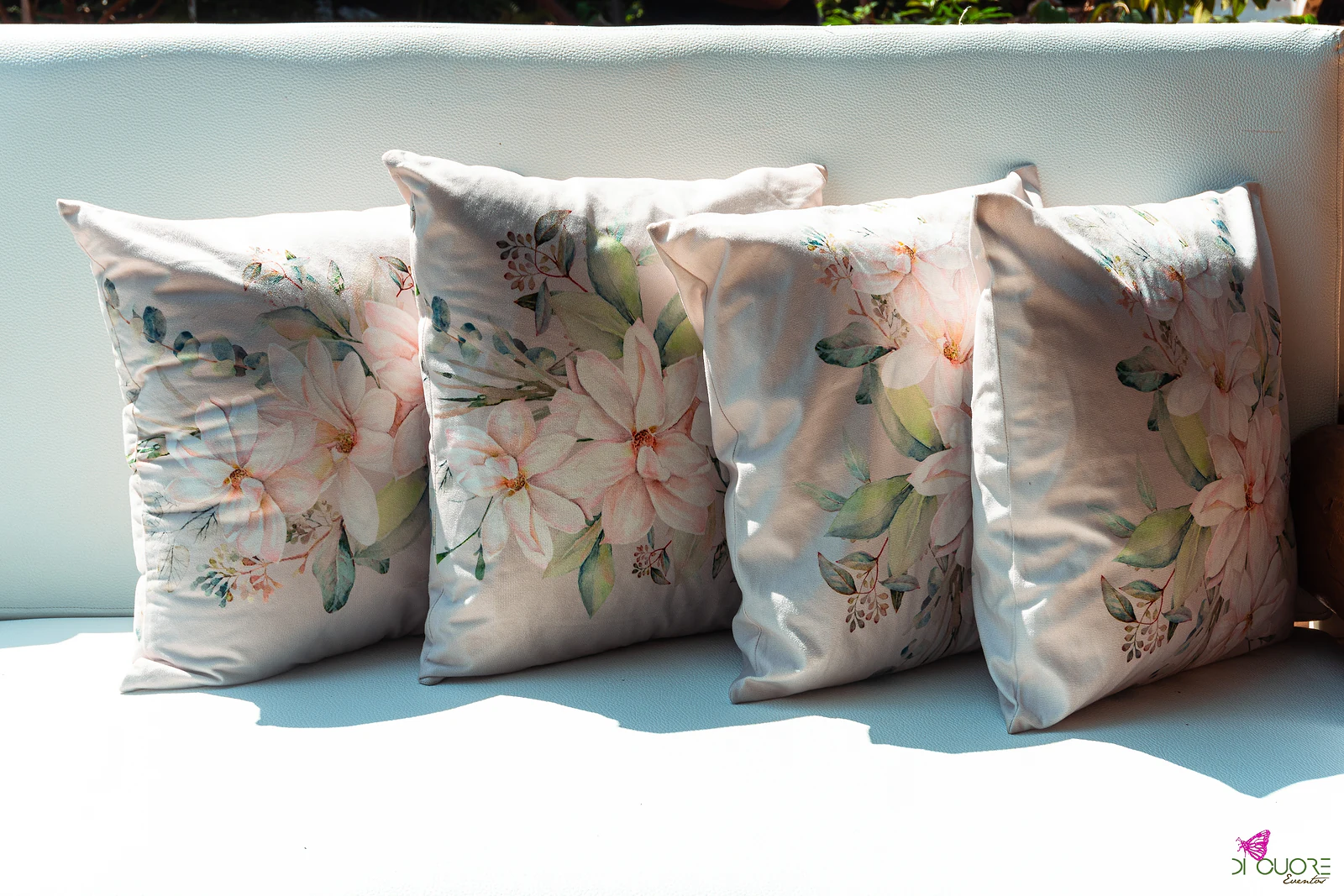 Quatro almofadas com flores pintadas em tons de rosa e folhas verdes, alinhadas num sofá branco ao sol