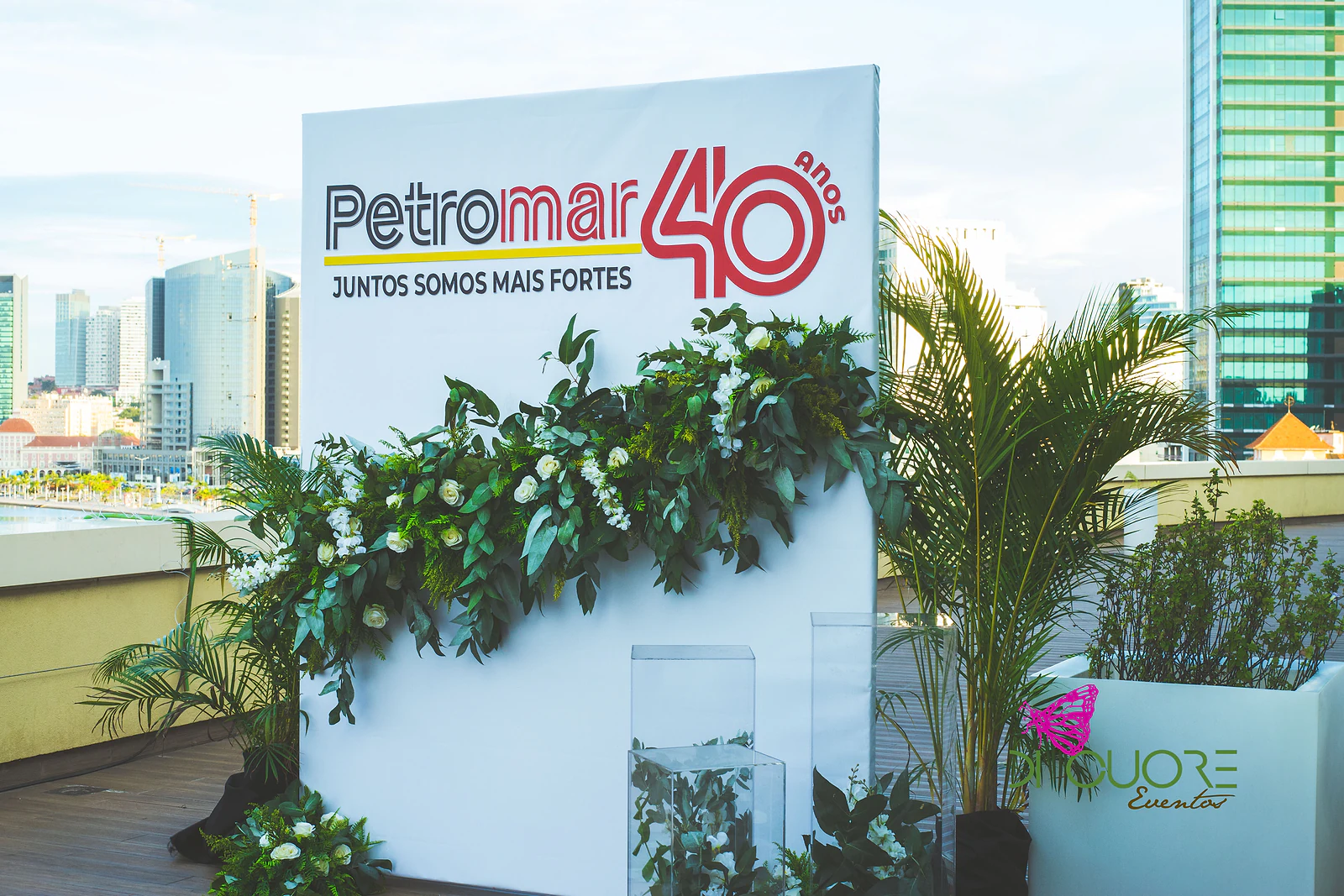 Painel branco com o logótipo «Petromar 40 anos – Juntos somos mais fortes», coberto por uma guirlanda de folhagem e flores brancas, num terraço com vista sobre a cidade