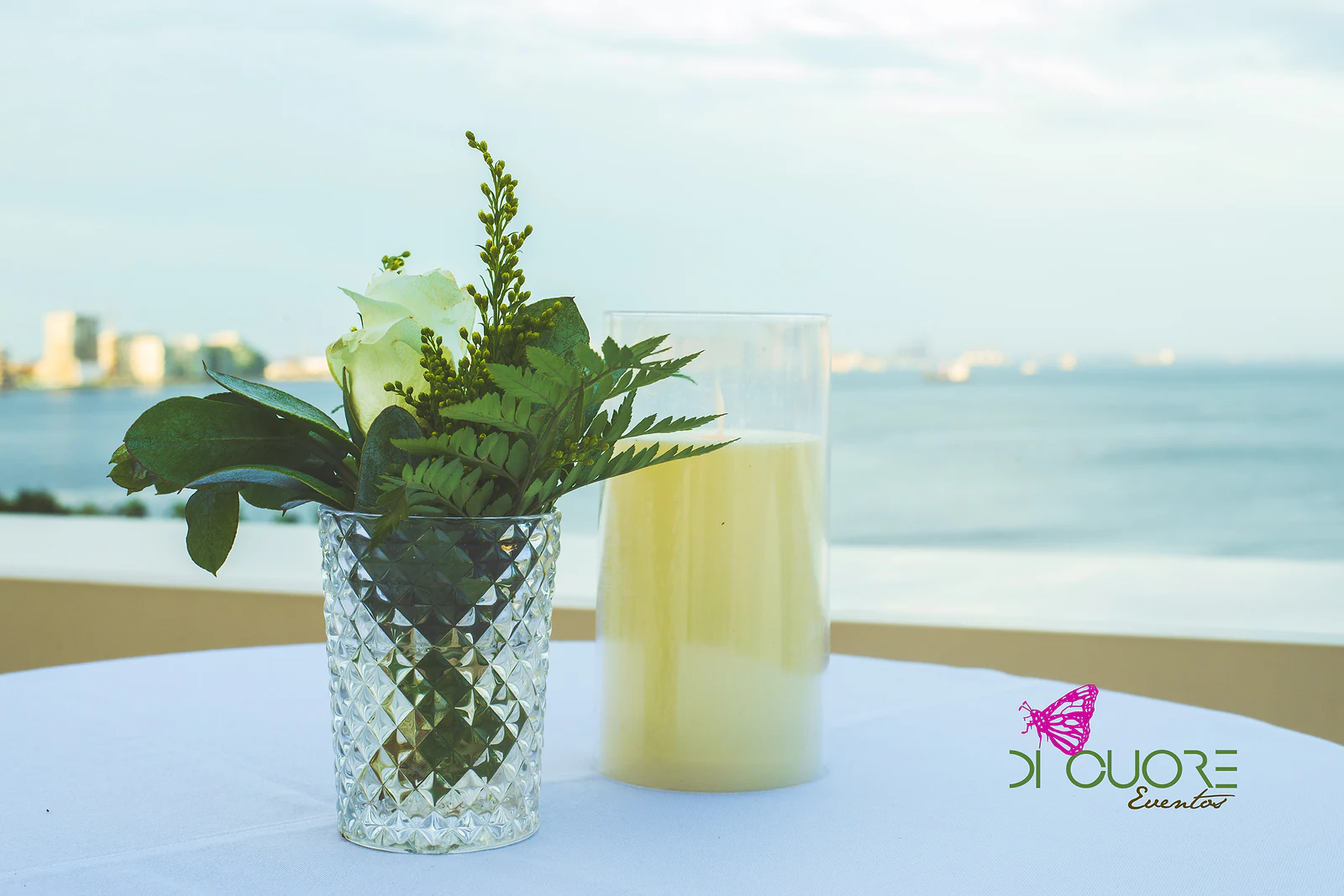 Copo de cristal lapidado com uma rosa branca e folhas de feto, ao lado de uma vela, sobre mesa de toalha branca com o mar ao fundo