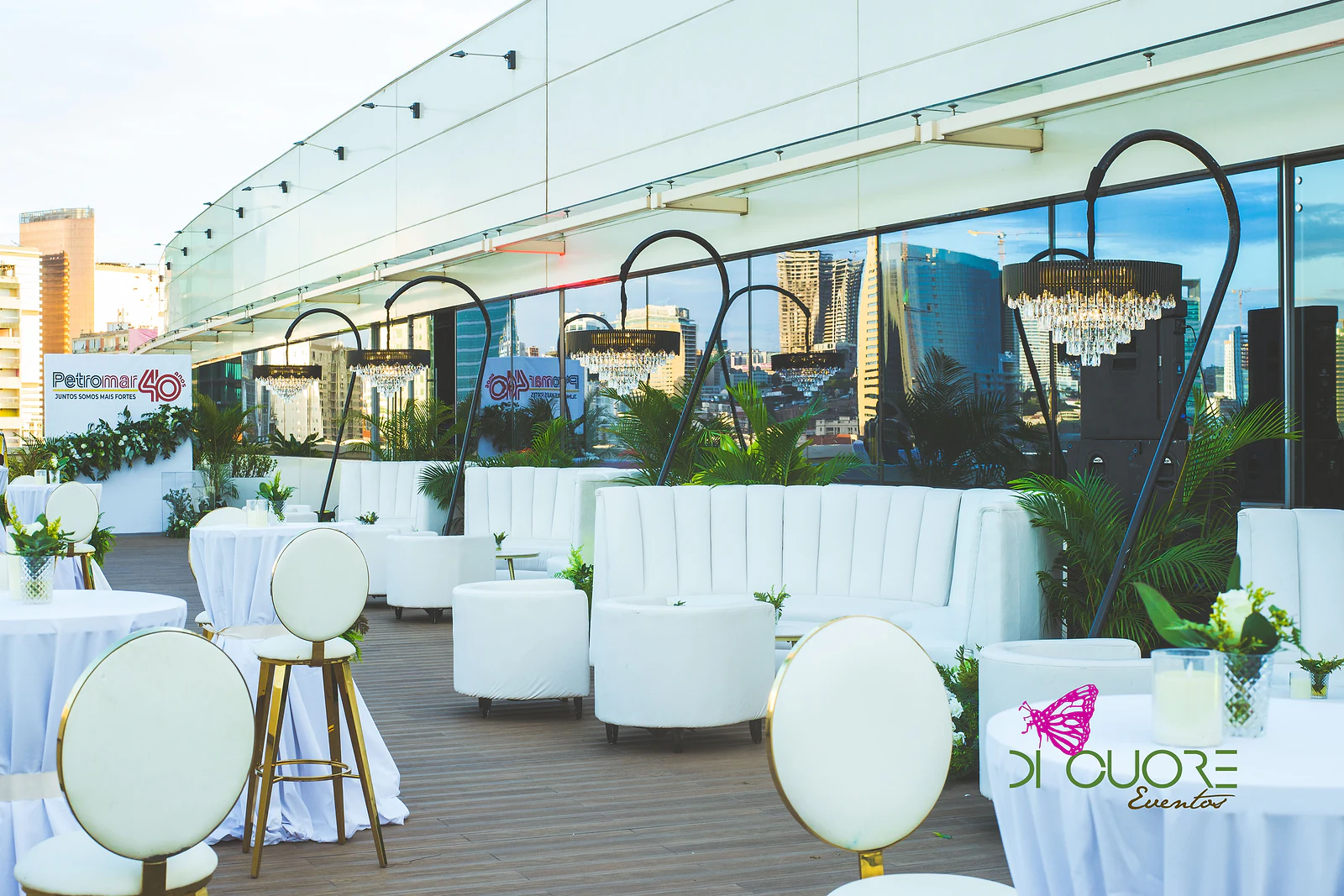 Terraço com mesas altas de saia branca e bancos de costas redondas, sofás brancos sob candeeiros de cristal e o painel Petromar 40 anos ao fundo