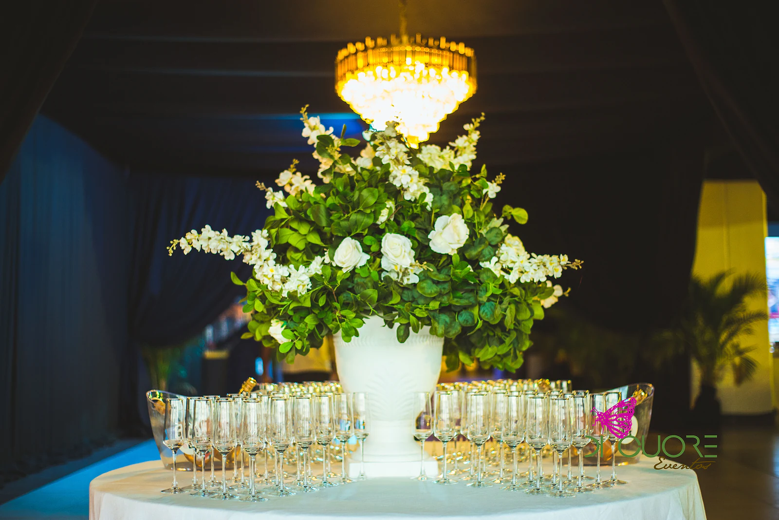 Grande vaso branco com folhagem e flores brancas sobre uma mesa cheia de taças, sob um lustre de cristal