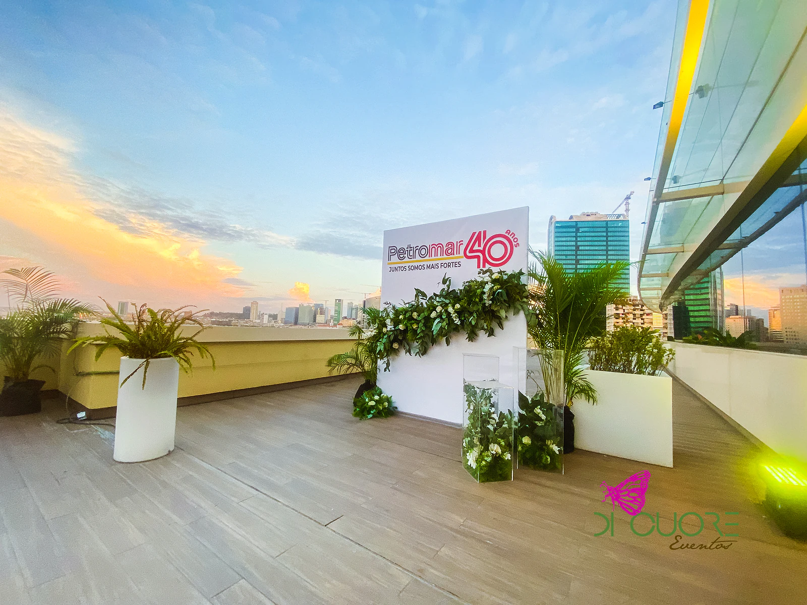 Terraço ao pôr do sol com o painel Petromar 40 anos, a grinalda de folhagem, plintos transparentes e vasos brancos com palmeiras, diante do horizonte da cidade