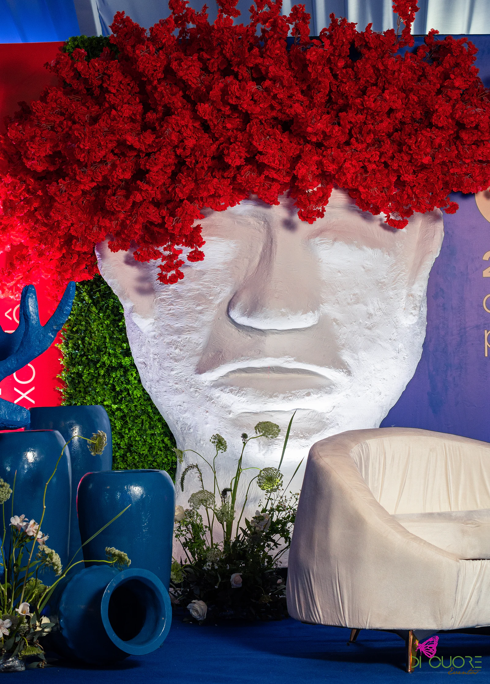Escultura branca de rosto com cabeleira de flores vermelhas, jarros azuis, folhagem e uma poltrona clara