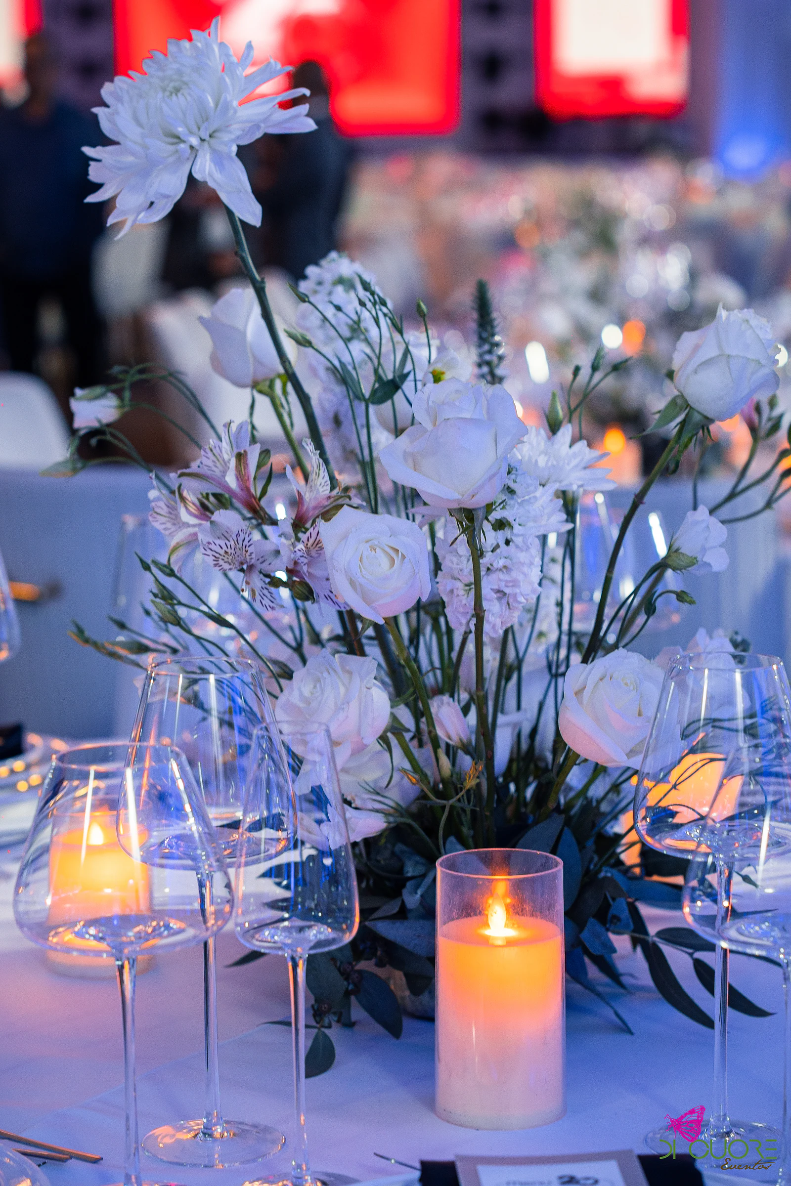 Rosas brancas, crisântemo e folhagem num centro de mesa entre copos de cristal e velas acesas, em luz azulada