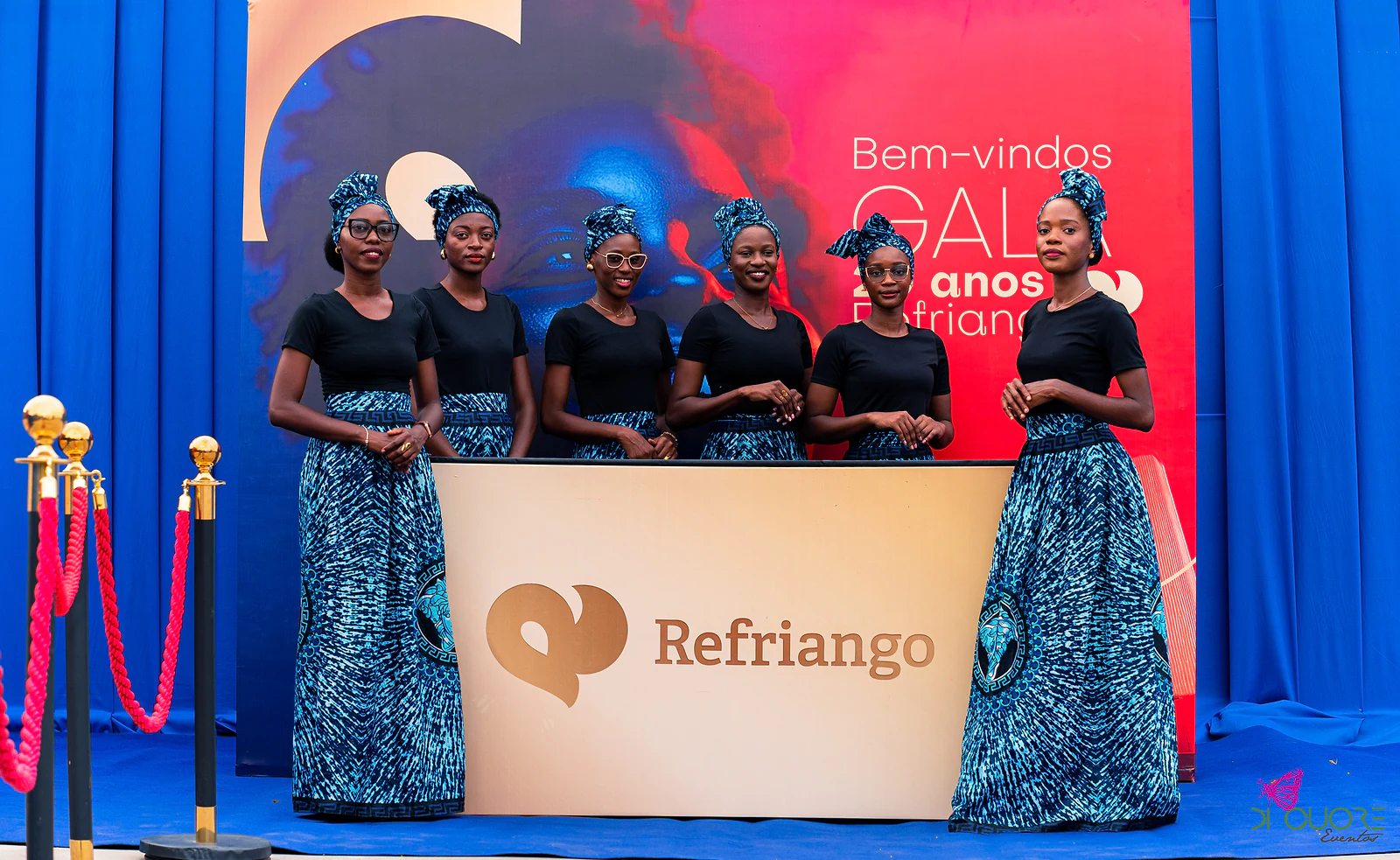 Seis anfitriãs de saias estampadas em azul e lenços na cabeça, atrás do balcão Refriango, diante do painel de boas-vindas da gala
